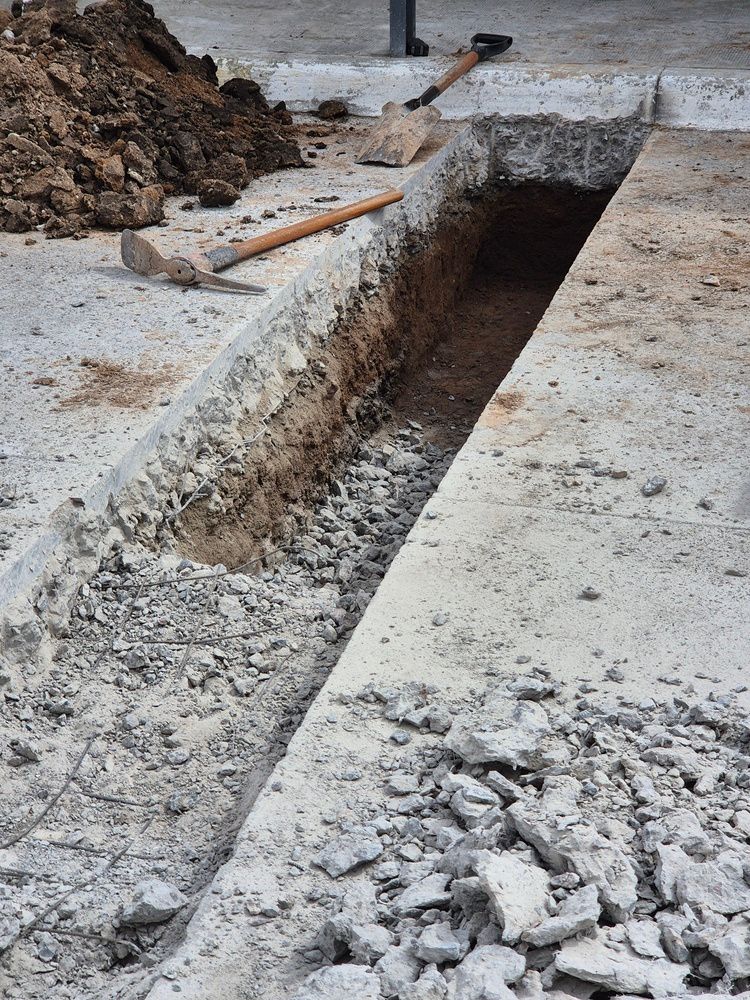 Trench dug in concrete. A pickaxe and shovel lie nearby, with a pile of dirt.