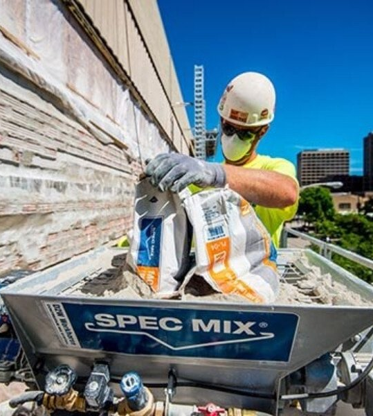 Construction worker mixing SPEC MIX mortar on a building exterior, sunny day.