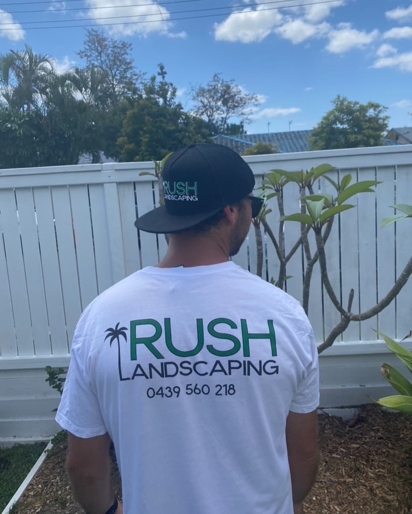 A man wearing a rush landscaping shirt is standing in front of a white fence