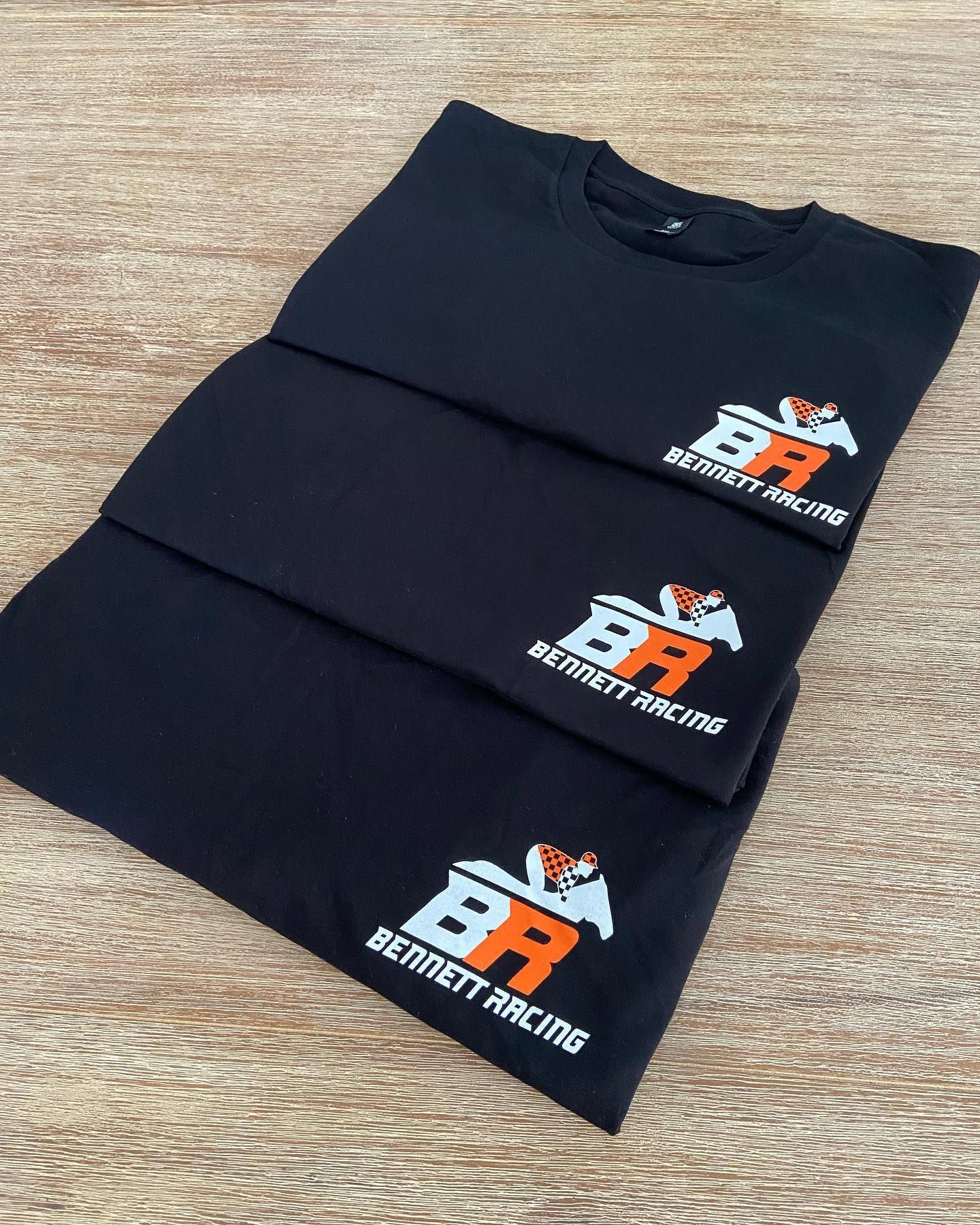 Three black t-shirts are stacked on top of each other on a wooden table.