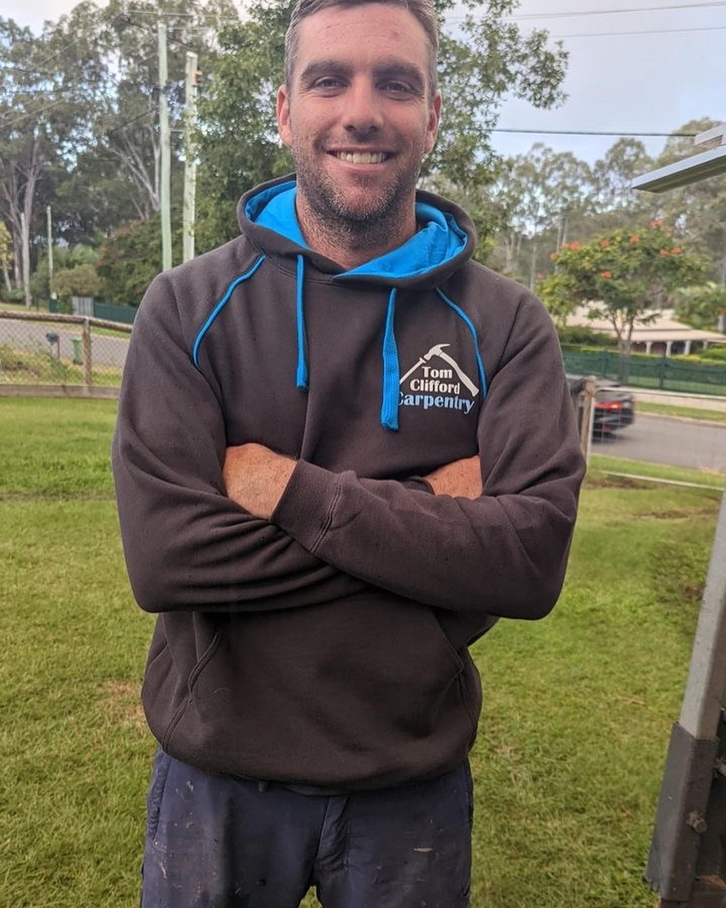 A man wearing a brown hoodie with a blue hood is standing in the grass with his arms crossed.