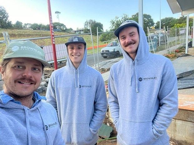 Three men wearing hoodies and hats are standing next to each other on a construction site.