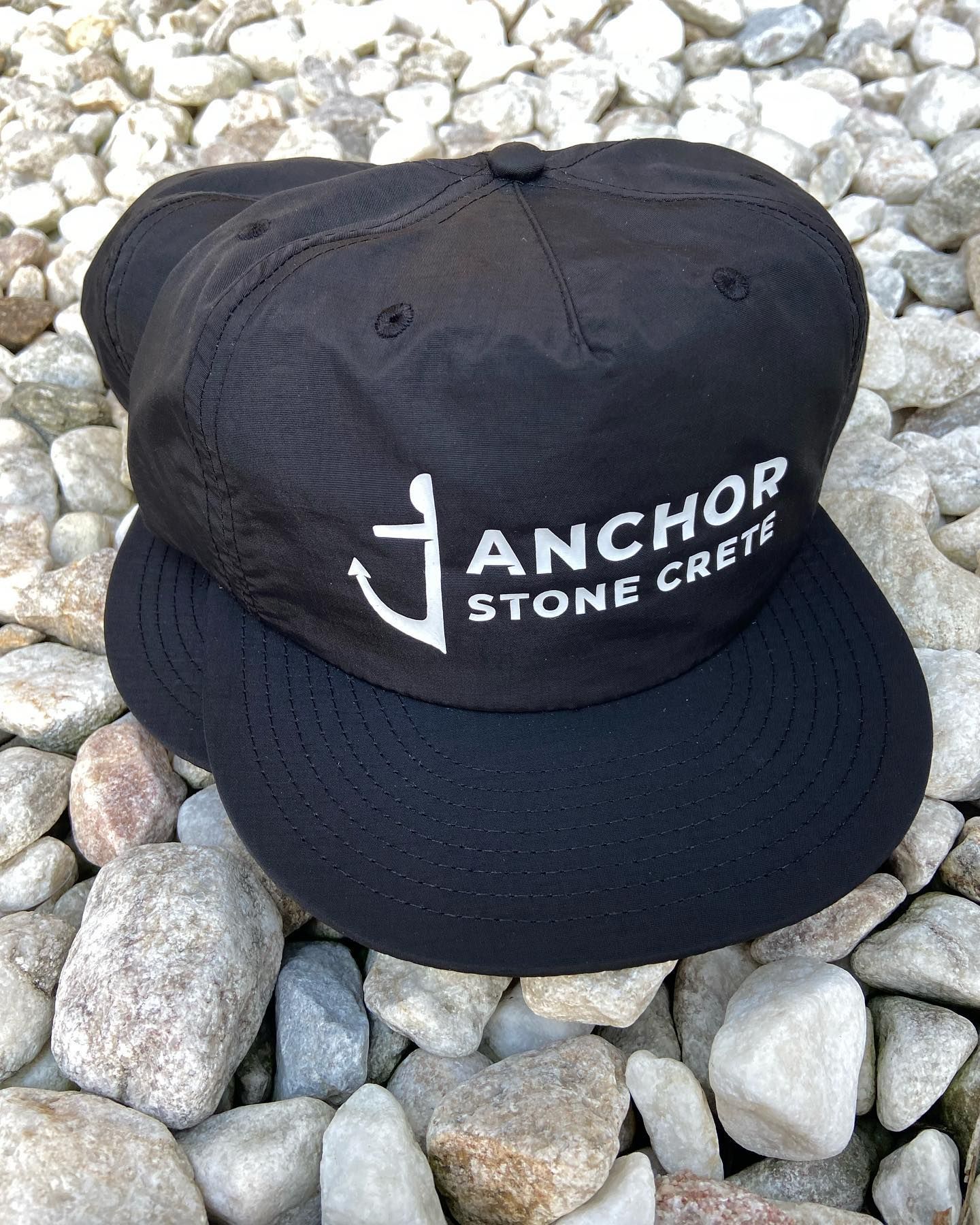 A black anchor stone crew hat is sitting on a pile of rocks.