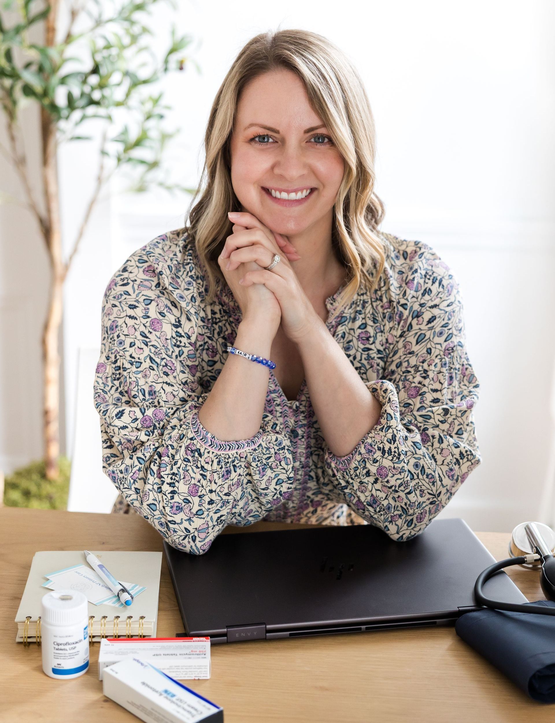 Woman with hands clasped, smiling at desk with laptop, medication, and blood pressure cuff.