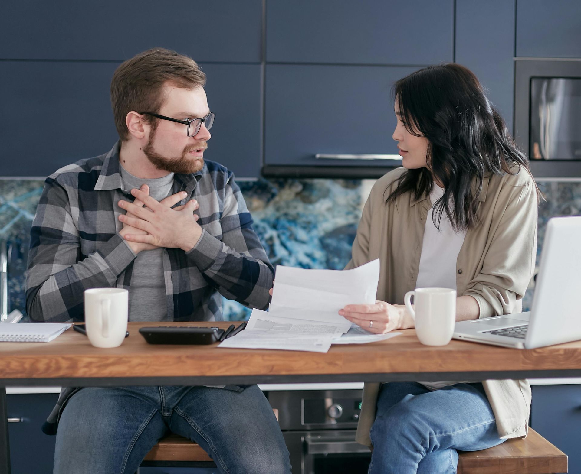 A person with a beard and glasses sits at a table with another person, reviewing documents with a laptop and mugs.