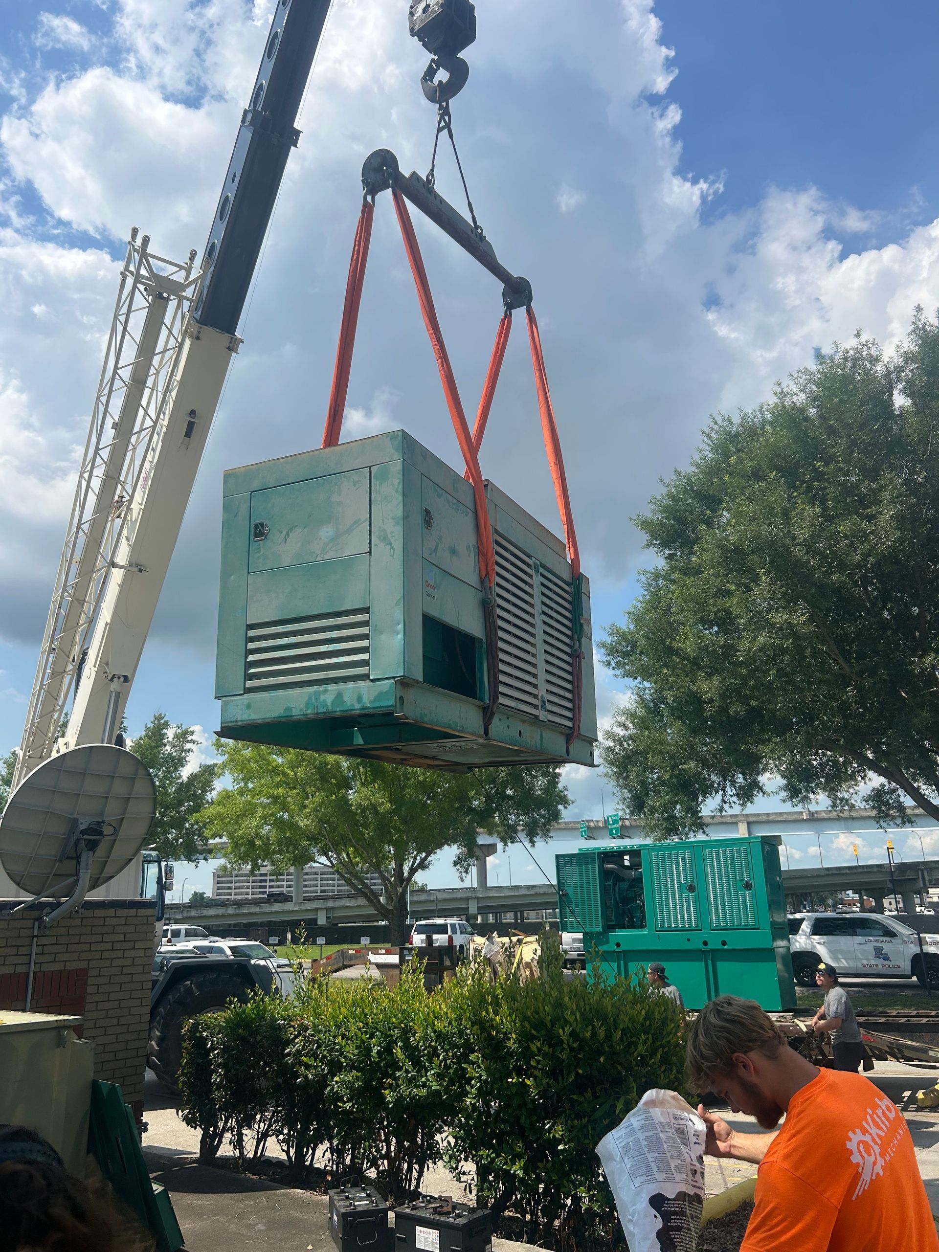 A crane lifting a large, green generator, with workers observing in an outdoor setting.