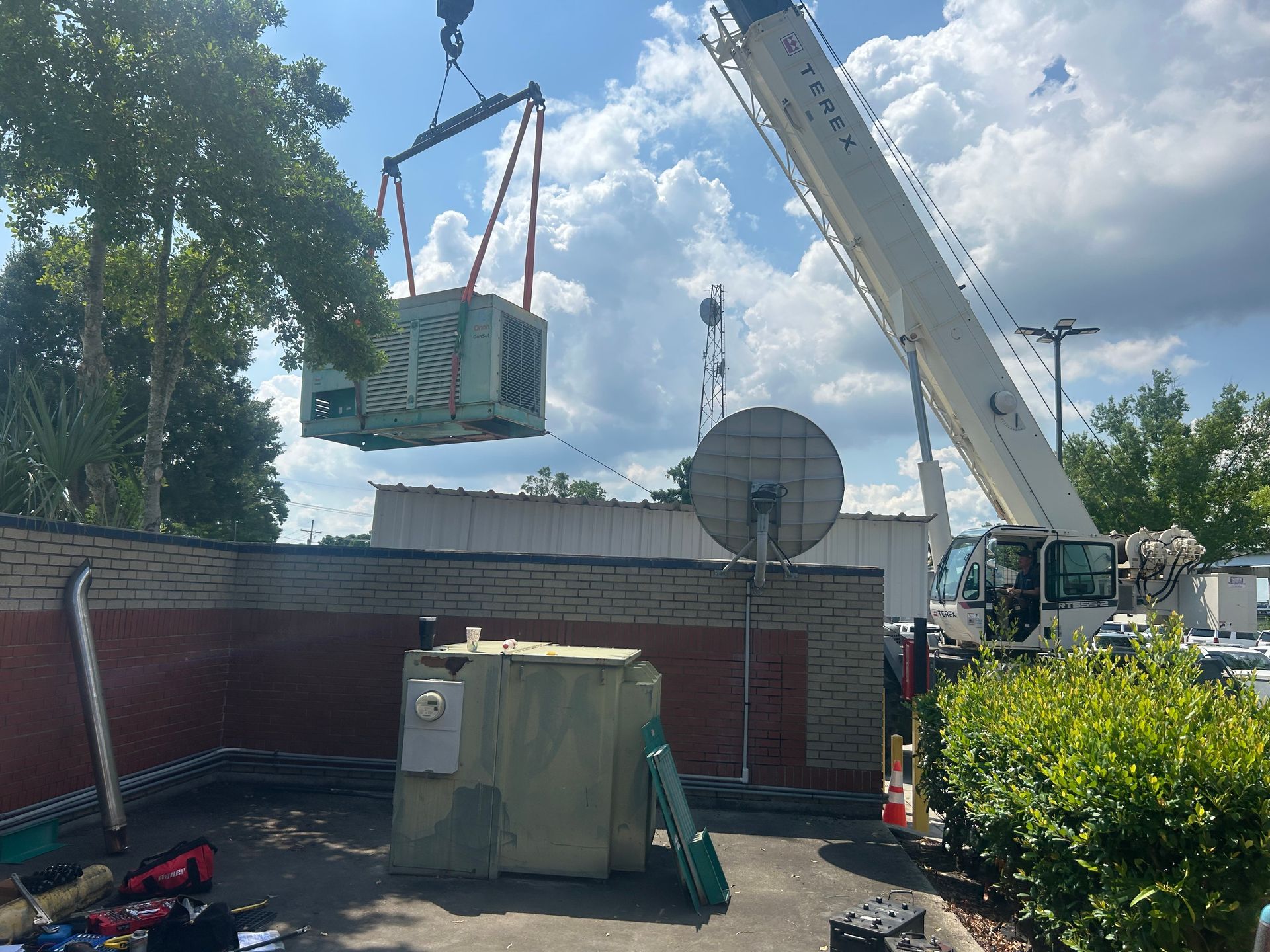 A crane lifting a large electrical transformer above a building with another on the ground. Sunny day.