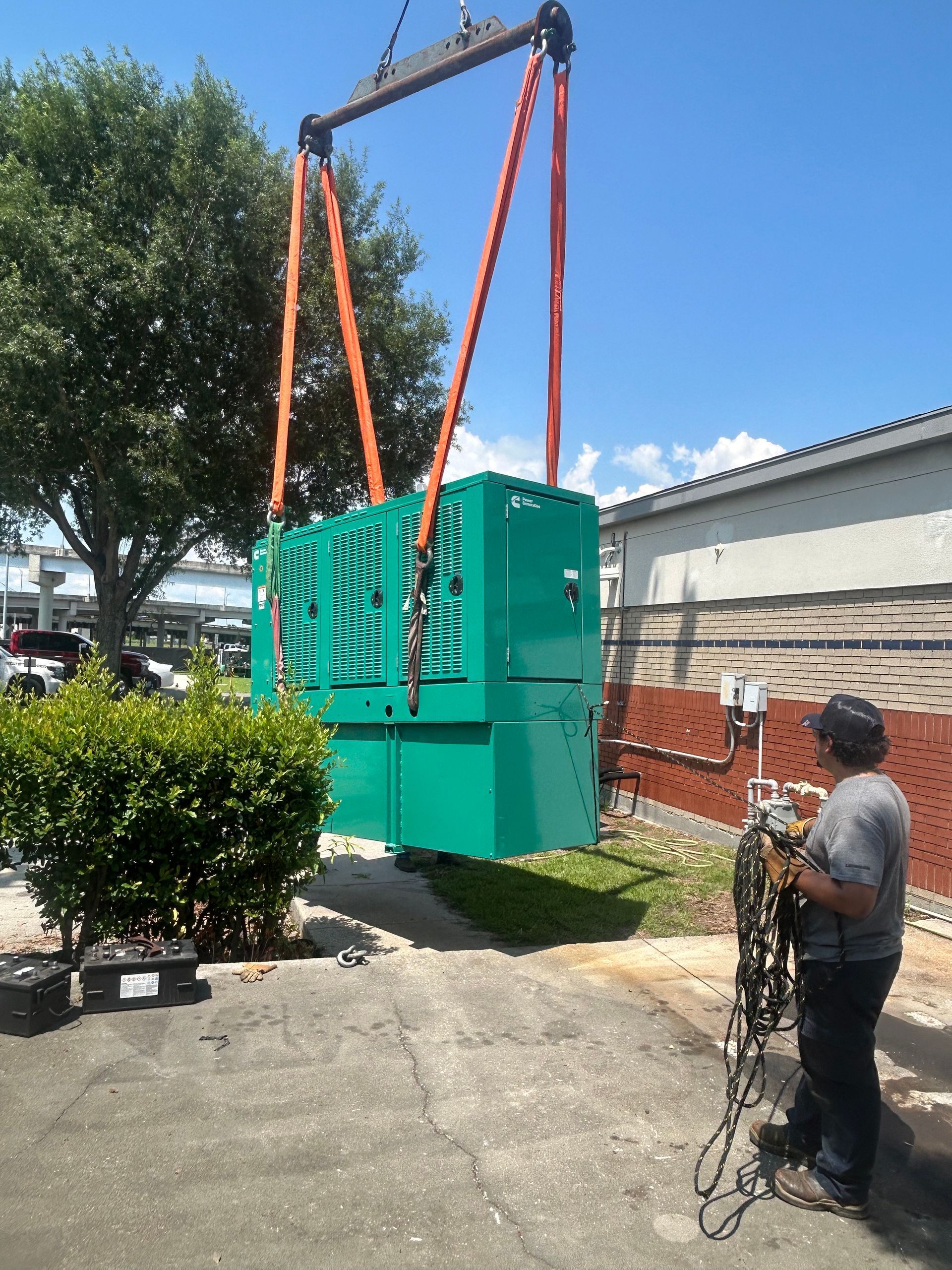 A large green generator is lifted by crane. A person watches, holding cables, outdoors on a sunny day.