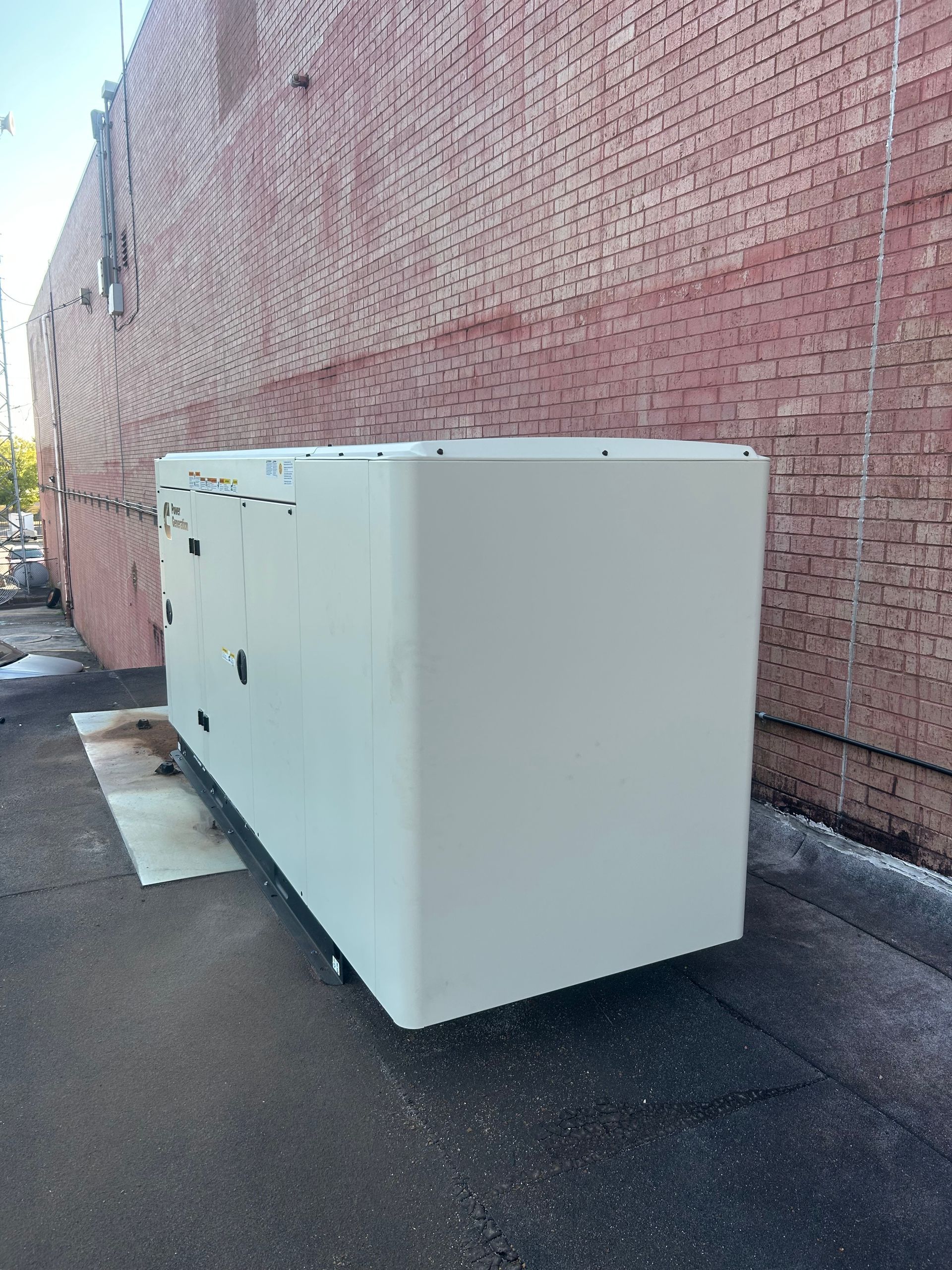 Large, white rectangular generator against a brick wall, on a paved surface.