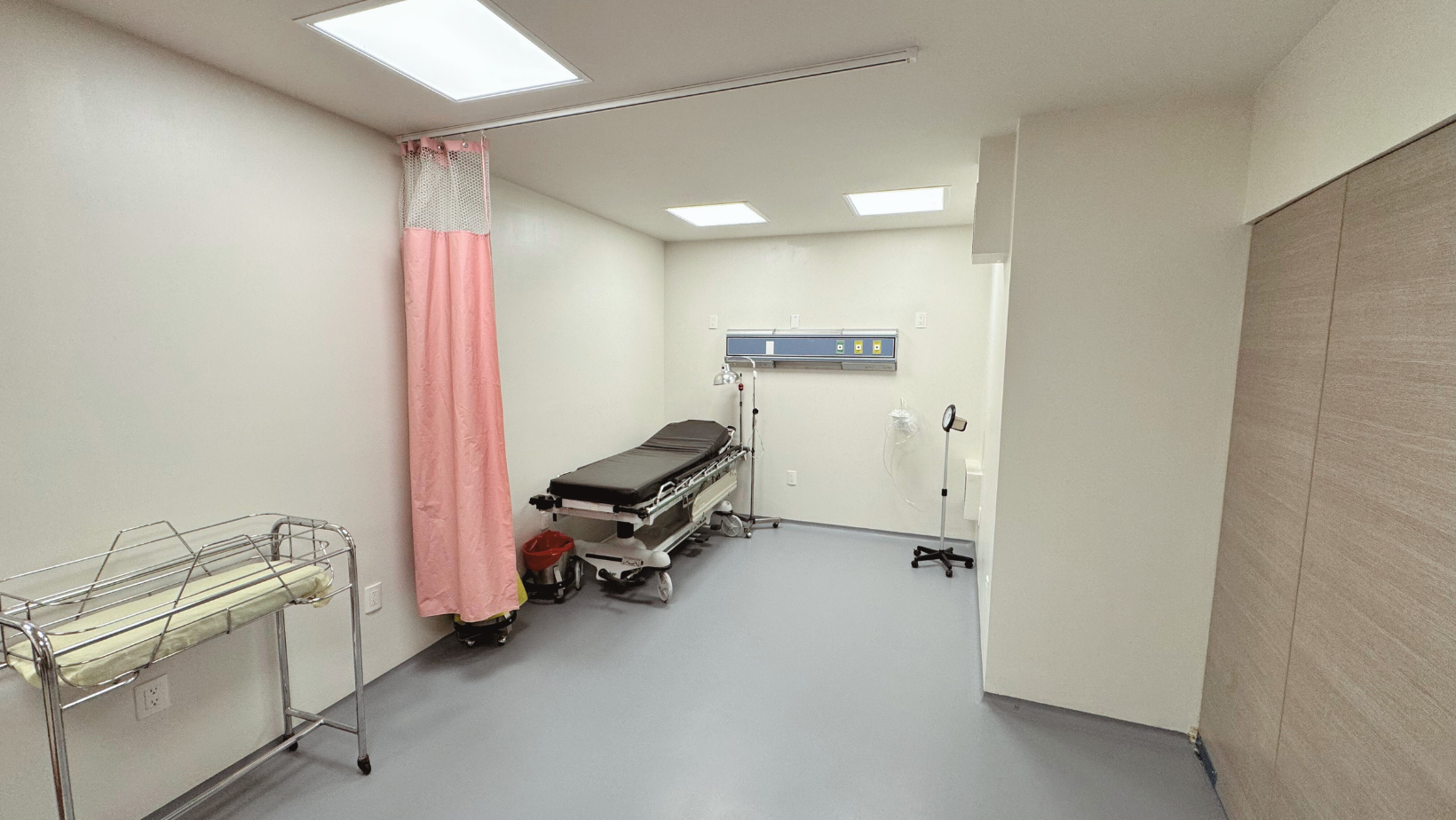 Sala de exploración: cama, cortina rosa, cambiador para bebés, equipo médico e iluminación brillante.