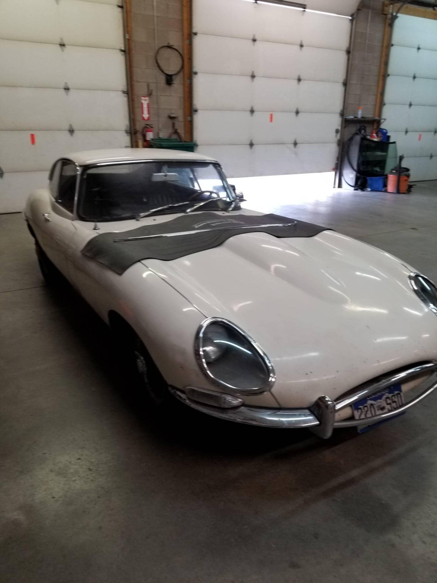 White Jaguar E-Type car inside a garage, covered with dark material on the hood, under bright lights.