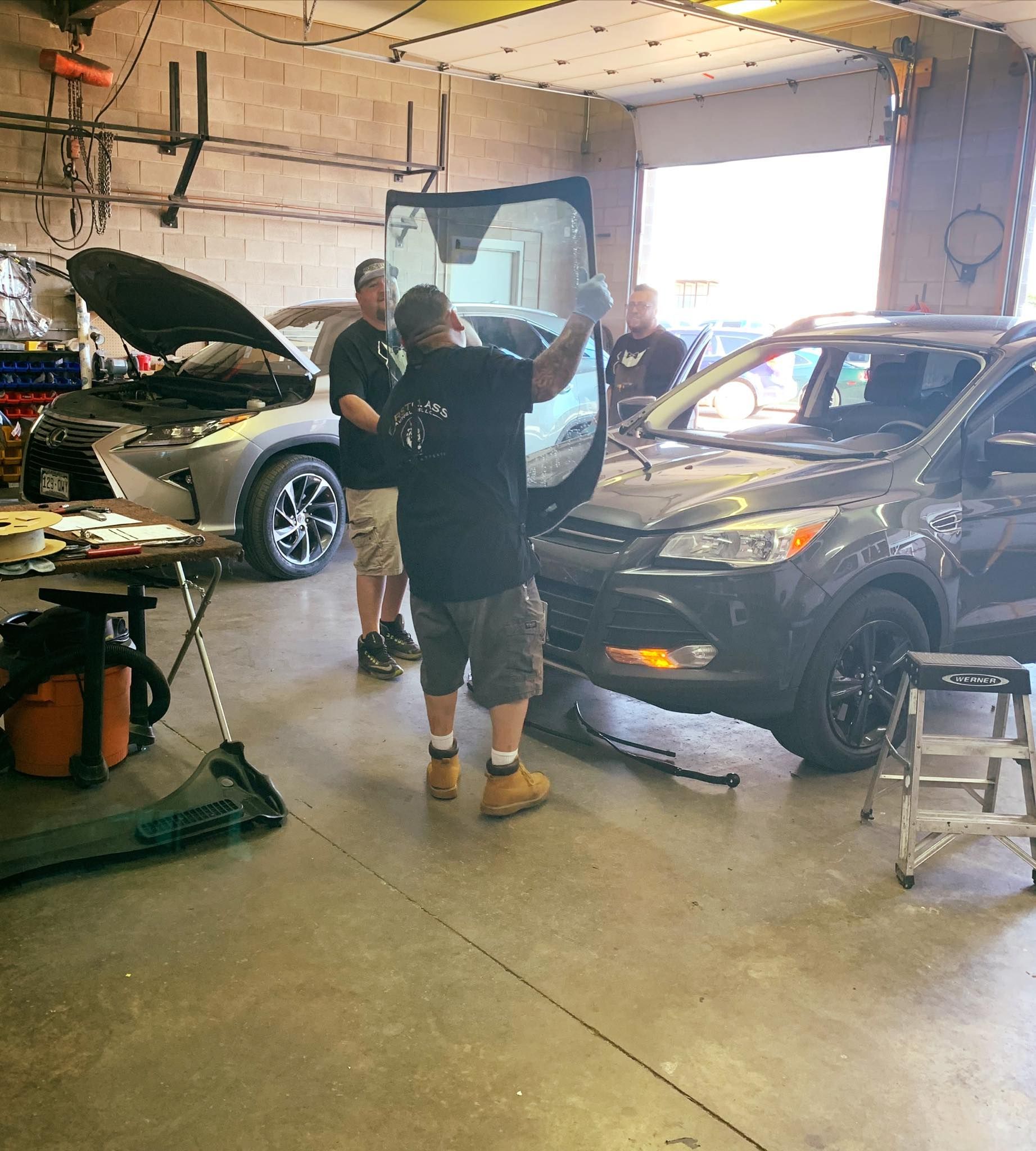 Three workers replacing a windshield on a grey SUV inside a garage. Another car with an open hood is in the background.