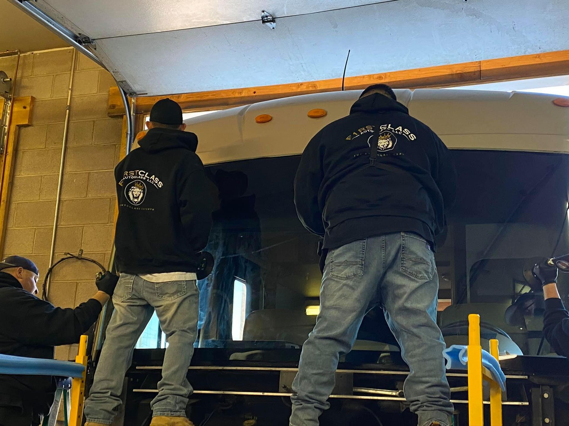 Two men in black hoodies lift a vehicle roof in a garage. Others assist.