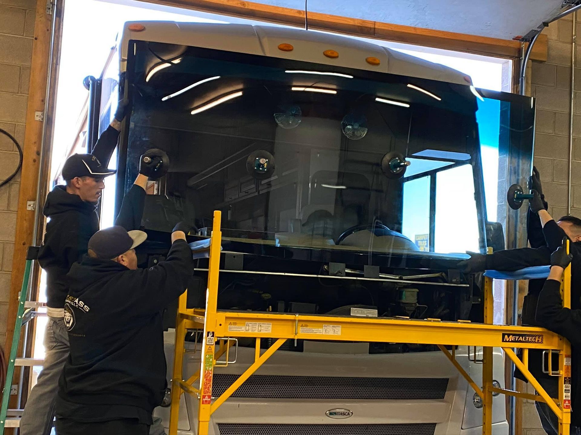 Four people replacing a large windshield on a vehicle inside a garage.