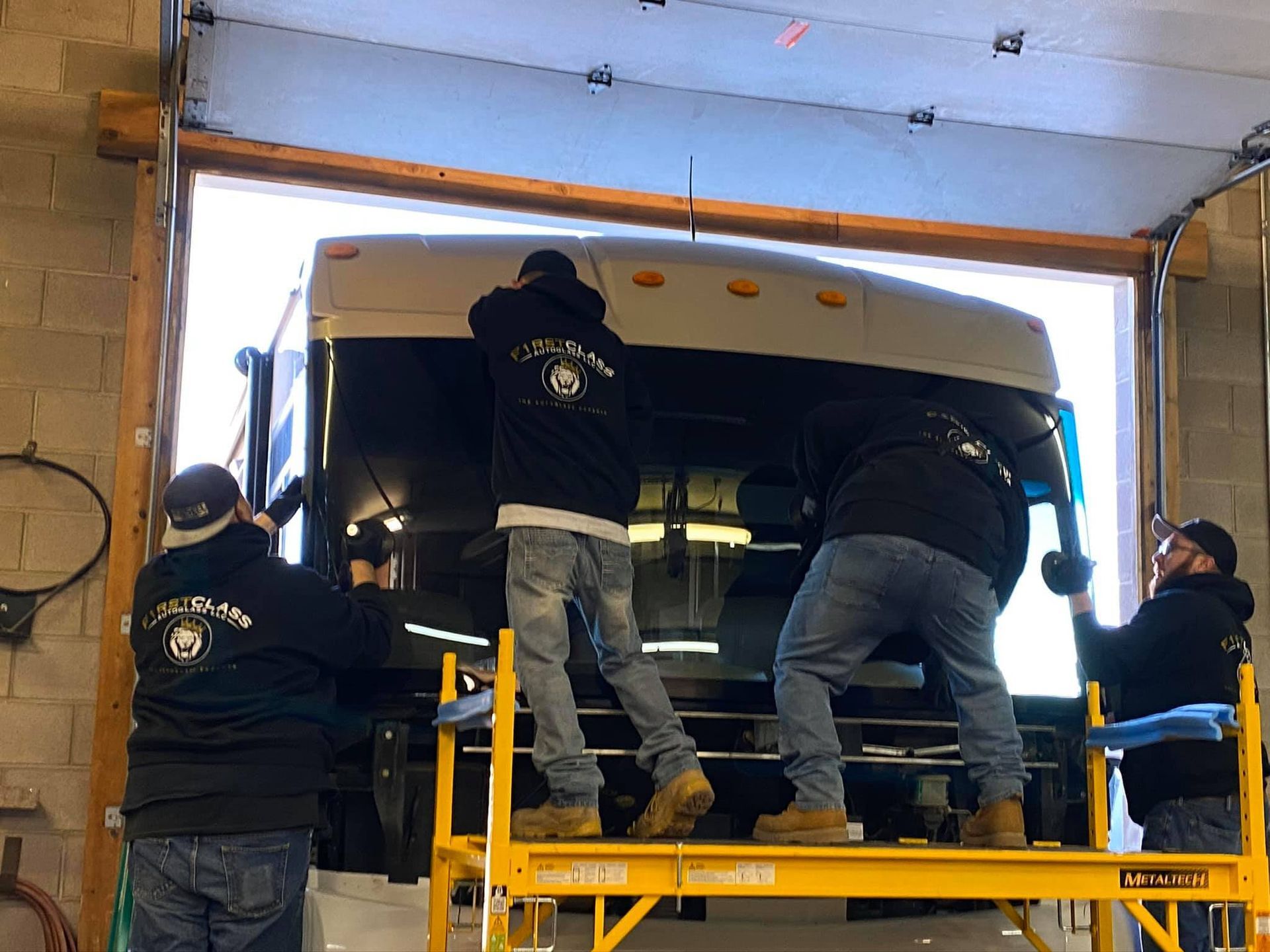 Four people installing part of a vehicle in an open garage doorway. They are on a yellow scaffolding.