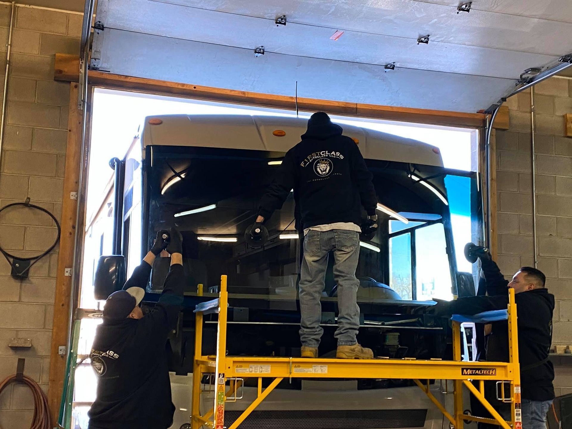 Workers installing a windshield on a bus in a garage, using scaffolding.