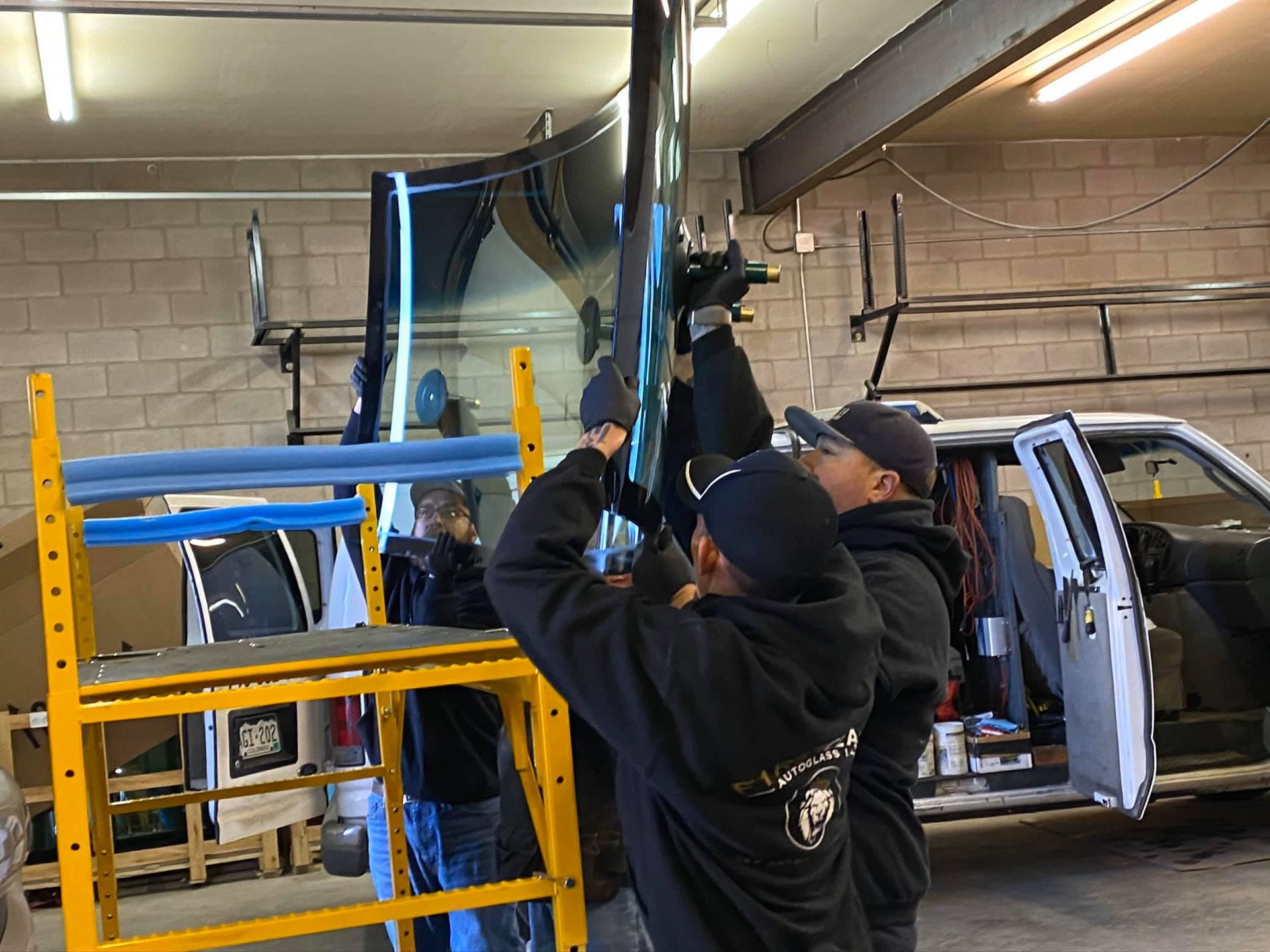 Three people installing a large tinted glass pane inside a workshop, near a yellow scaffold and a white truck.