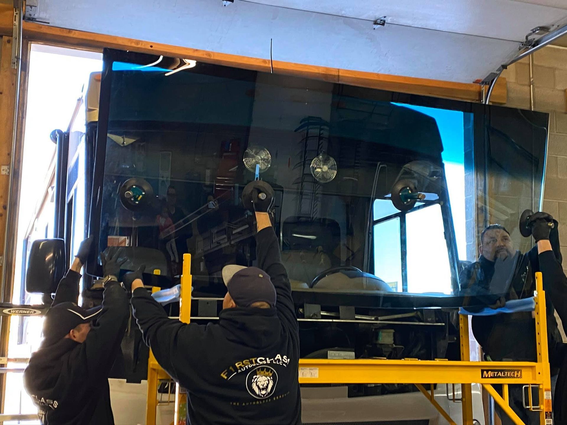 Three people installing a large, tinted windshield on a vehicle inside a garage.
