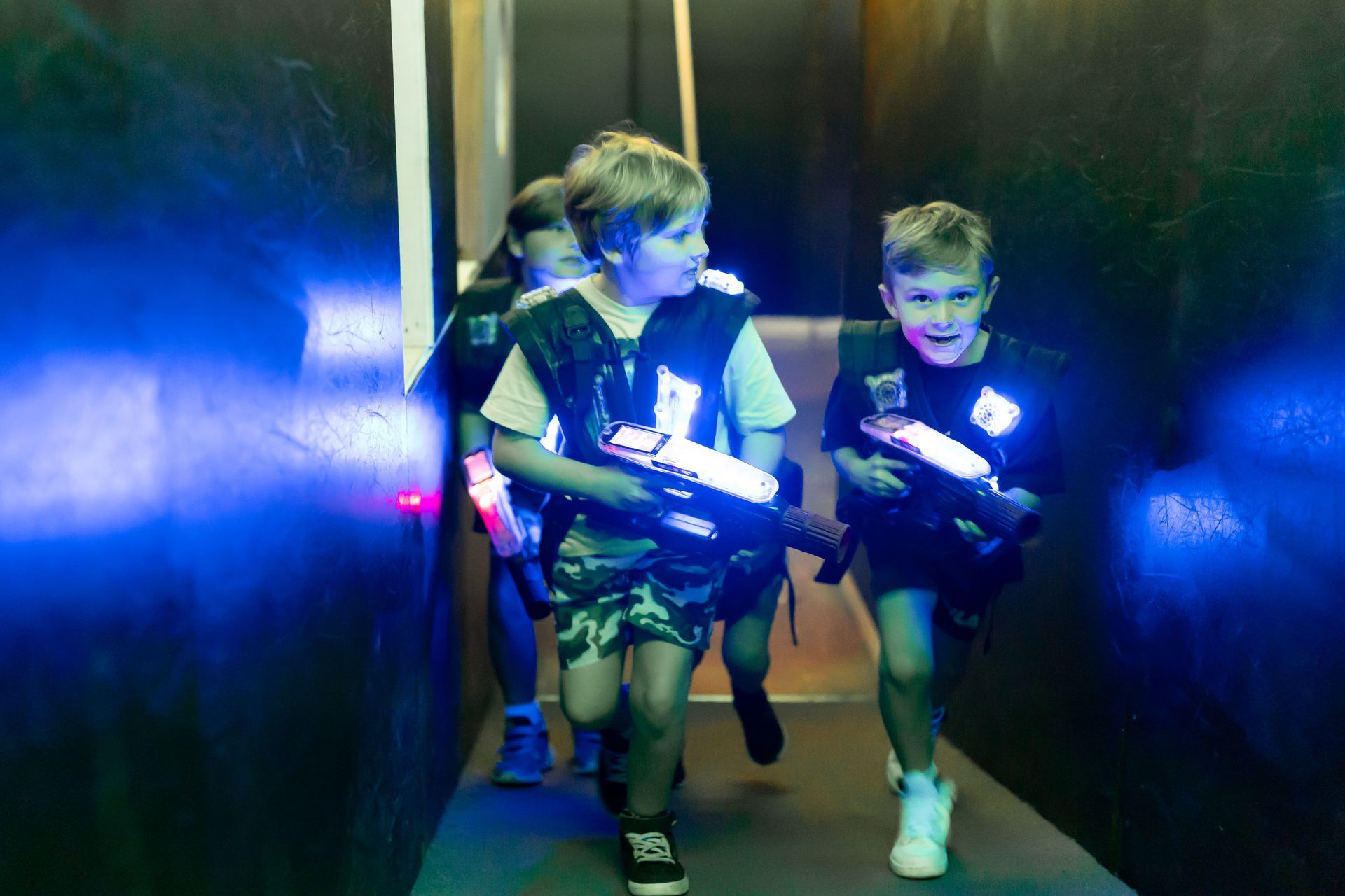 Three children moving through a laser tag arena, holding laser guns, with blue lights, at Laserzone Sunshine Coast.