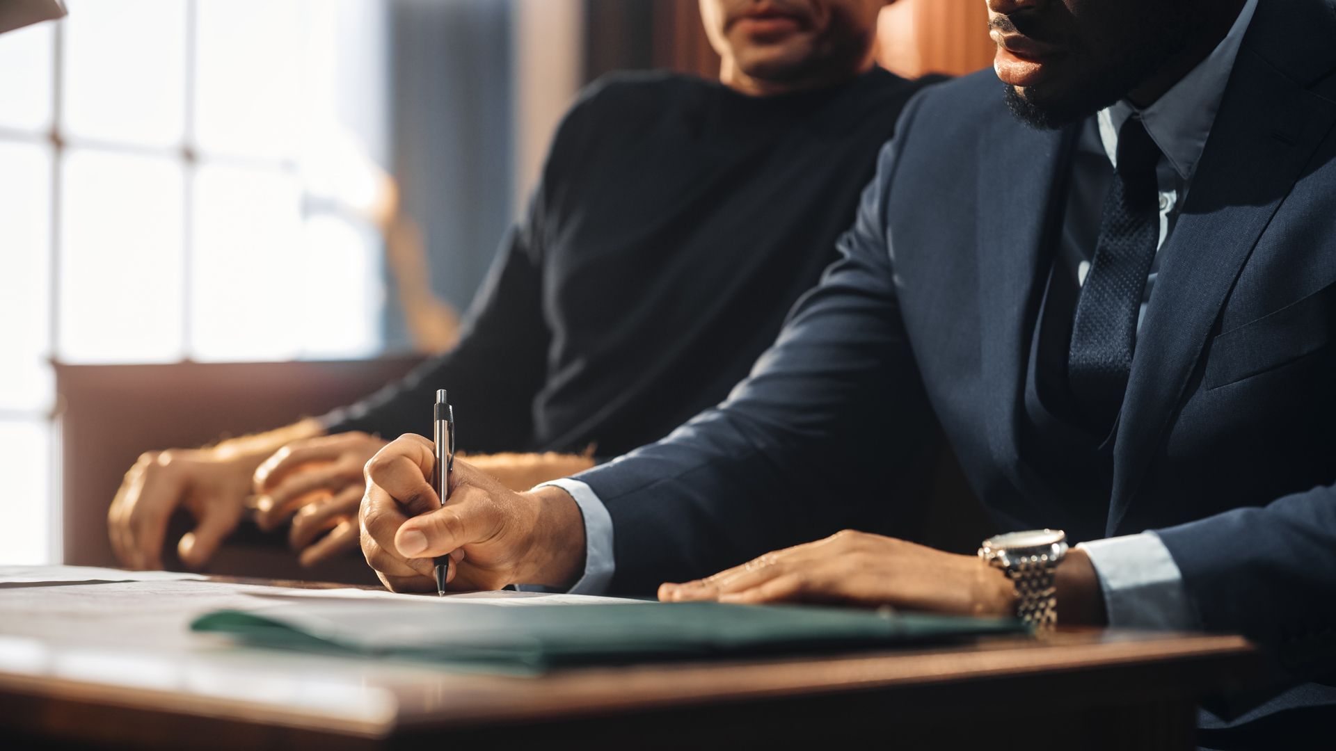 Two men in suits are sitting at a table signing a document
