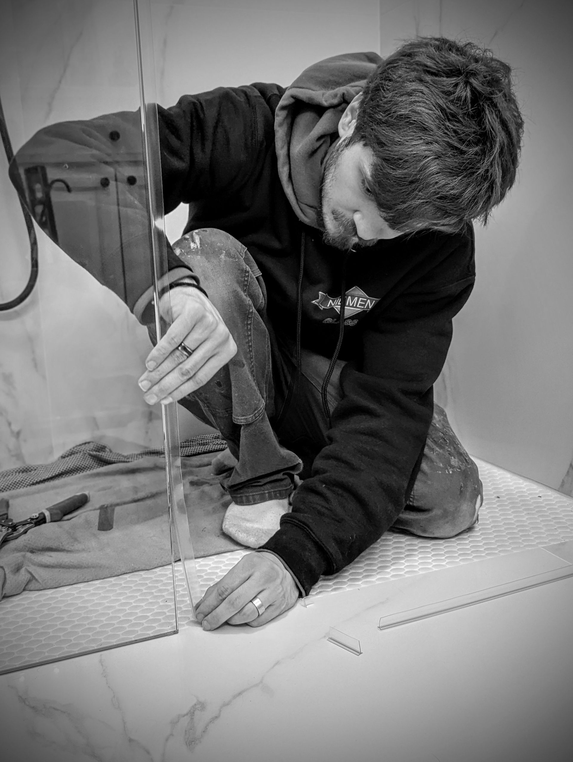 Man installing tile in a bathroom. He kneels, using a tool on the floor near a shower wall. Black and white.