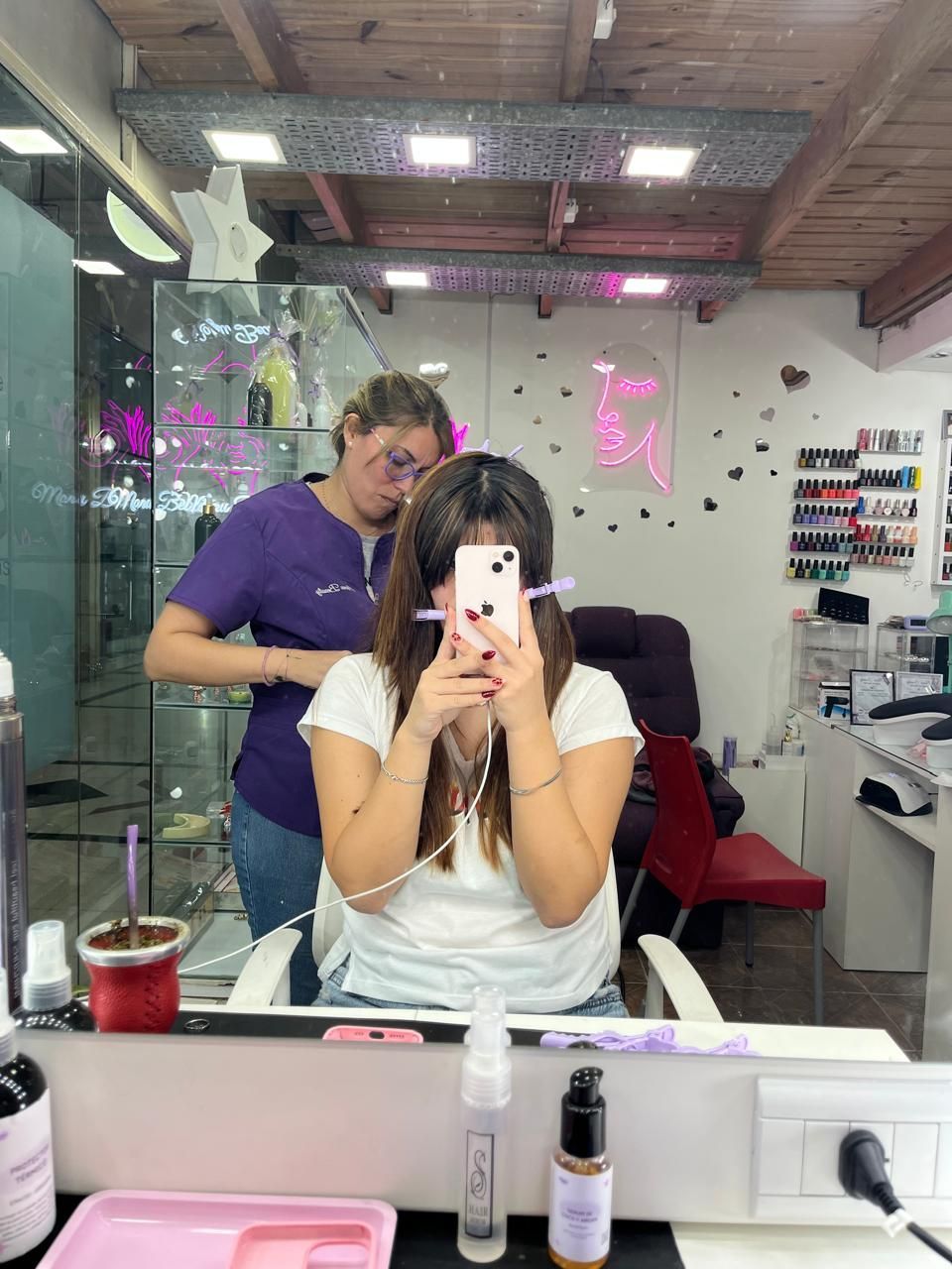 Una persona se toma una selfie frente al espejo en un salón de belleza mientras otra persona se encuentra detrás.
