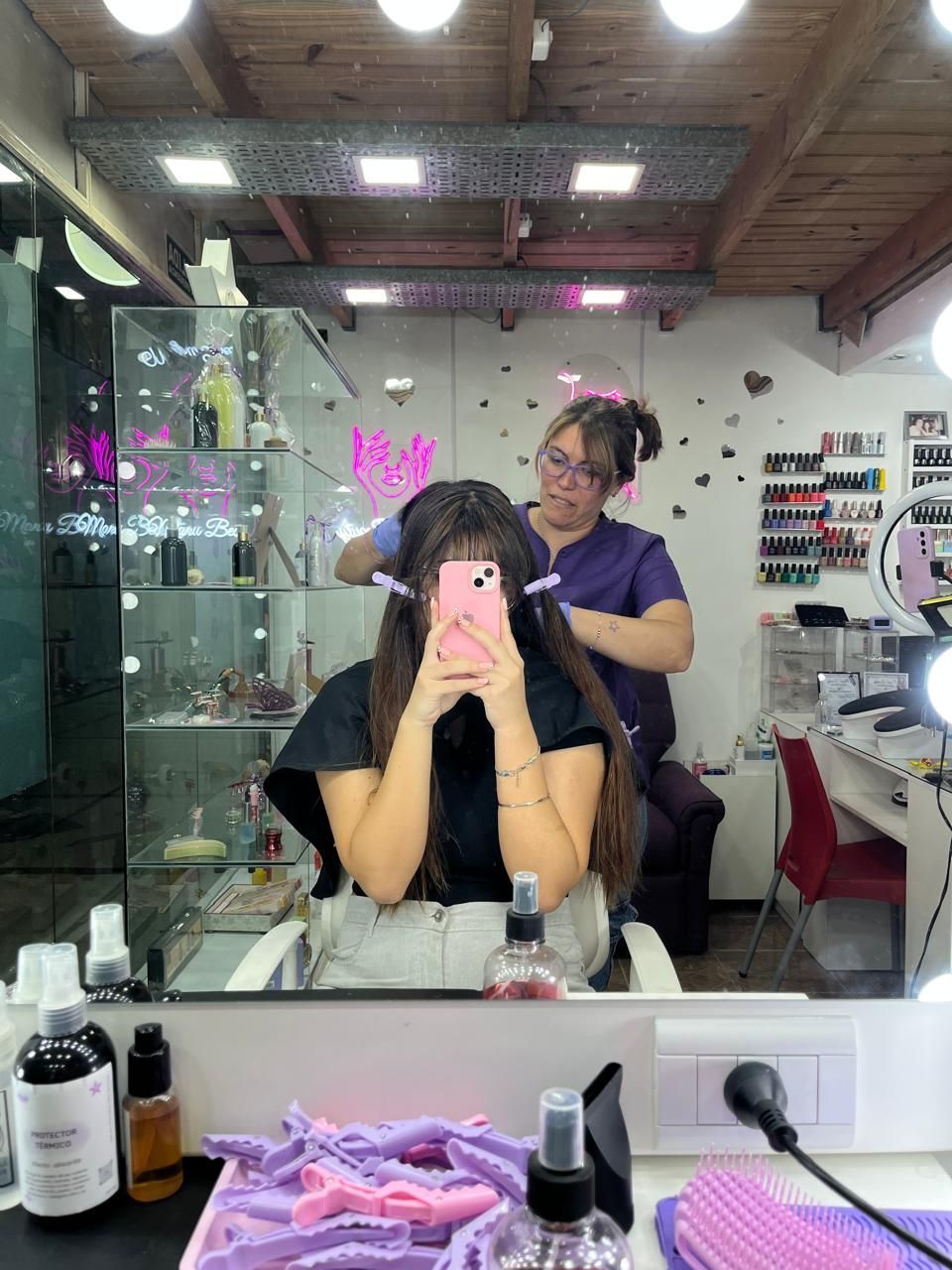 Una mujer se toma una selfie frente al espejo en una peluquería mientras una estilista se encuentra detrás de ella.