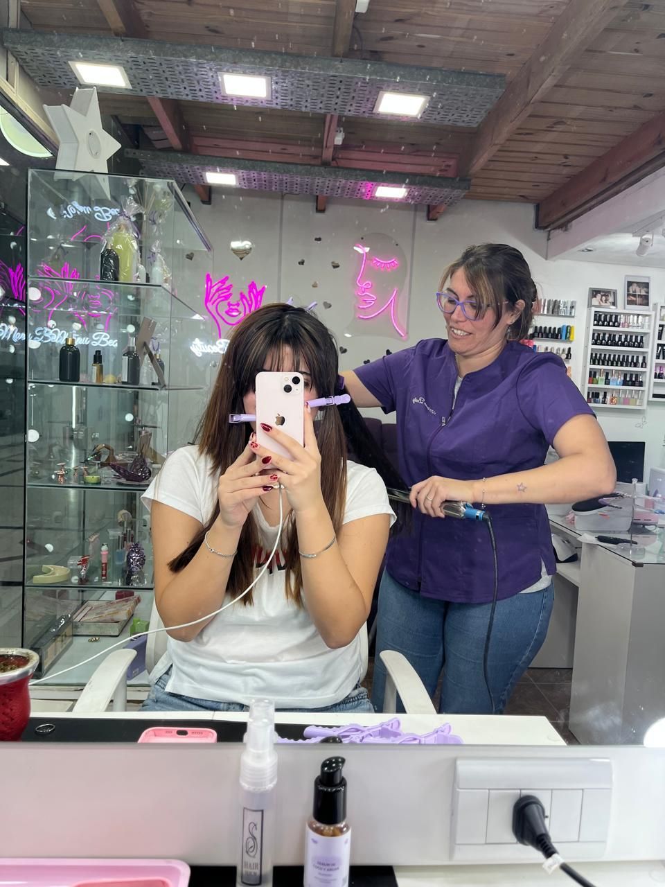 Una persona se toma una selfie frente al espejo en una peluquería mientras otra estilista se encuentra detrás.