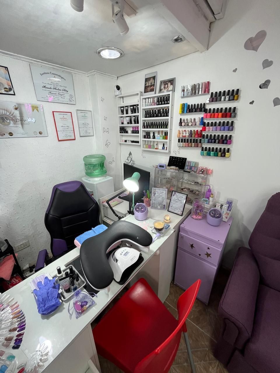 Pequeño salón de uñas con mesa de manicura, silla morada, silla de pedicura roja, estantes para esmaltes y paredes blancas.