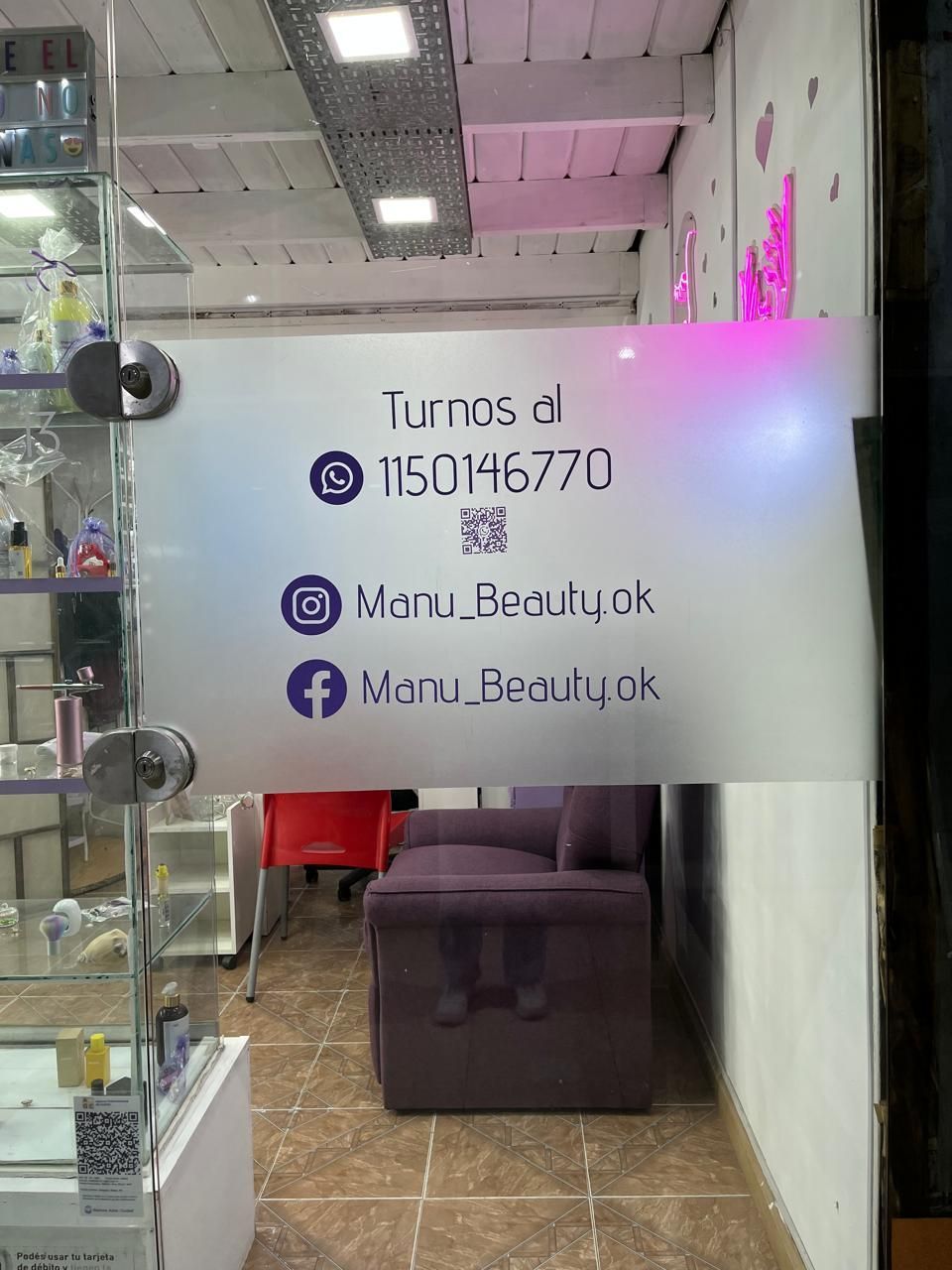 Pequeño local de peluquería con puerta de cristal esmerilado, iluminación rosa y cartel con información de contacto.