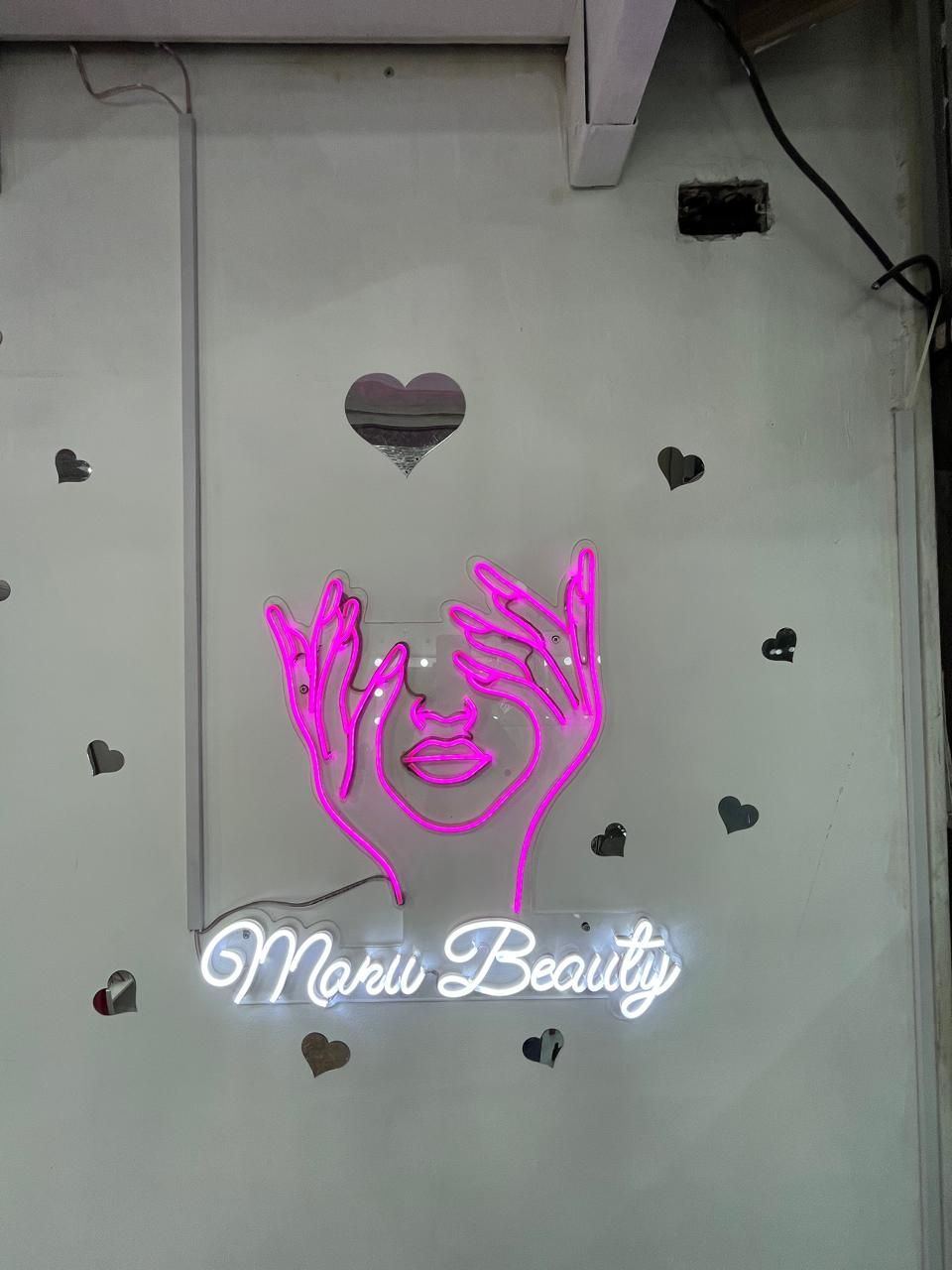 Letrero de neón con manos rosas alrededor de una cara, con “Mavi Beauty” brillando debajo en una pared decorada.