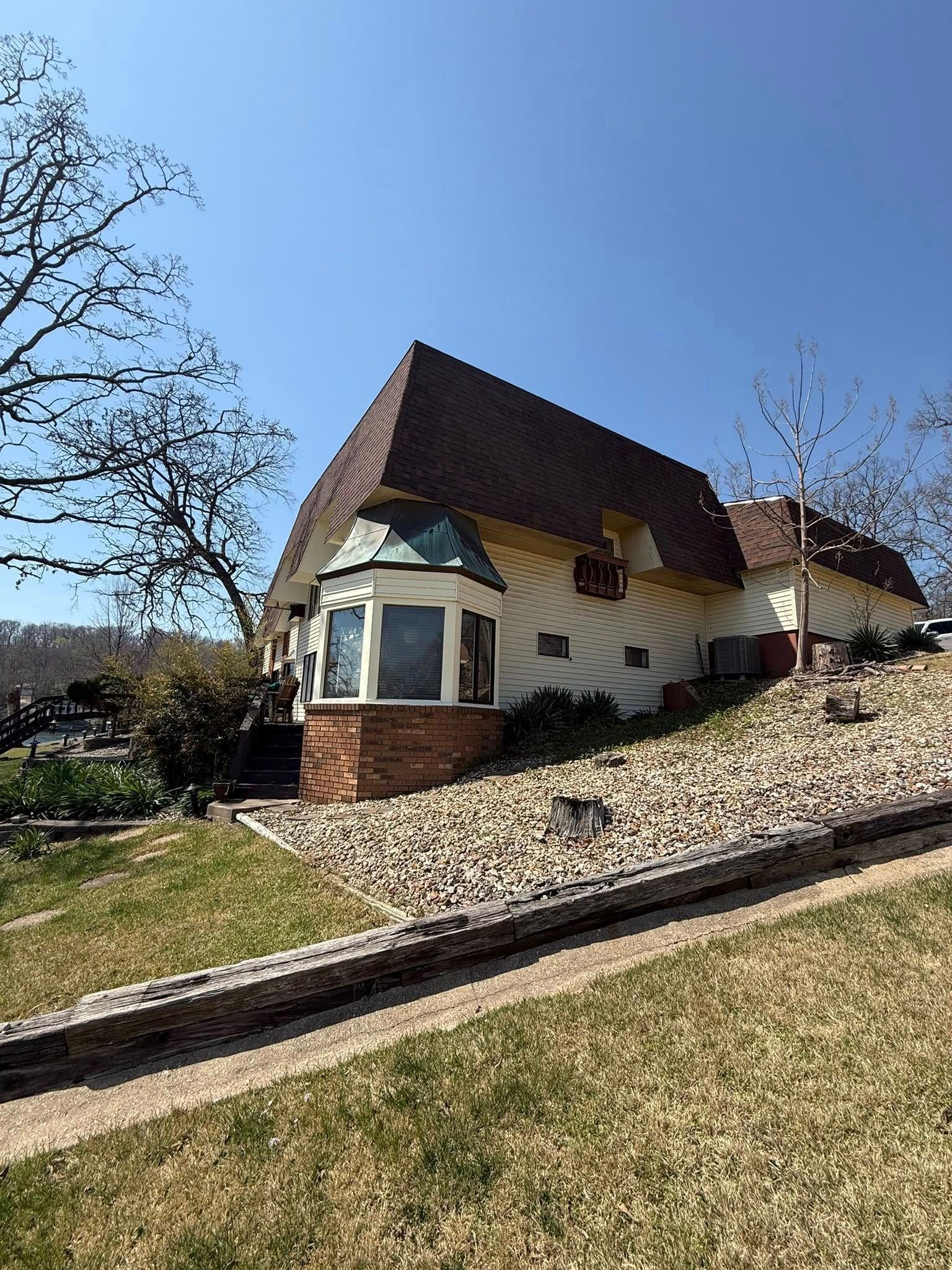 Whimsical house with angled brown roof, bay window, and beige siding on a hillside, under a bright blue sky.