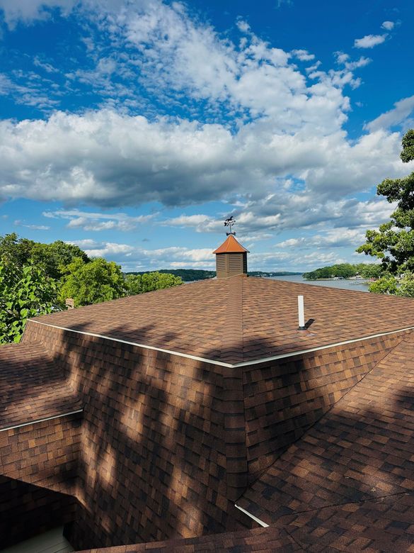 Brown shingled roof with a small cupola, tree shadows, and a partly cloudy blue sky.