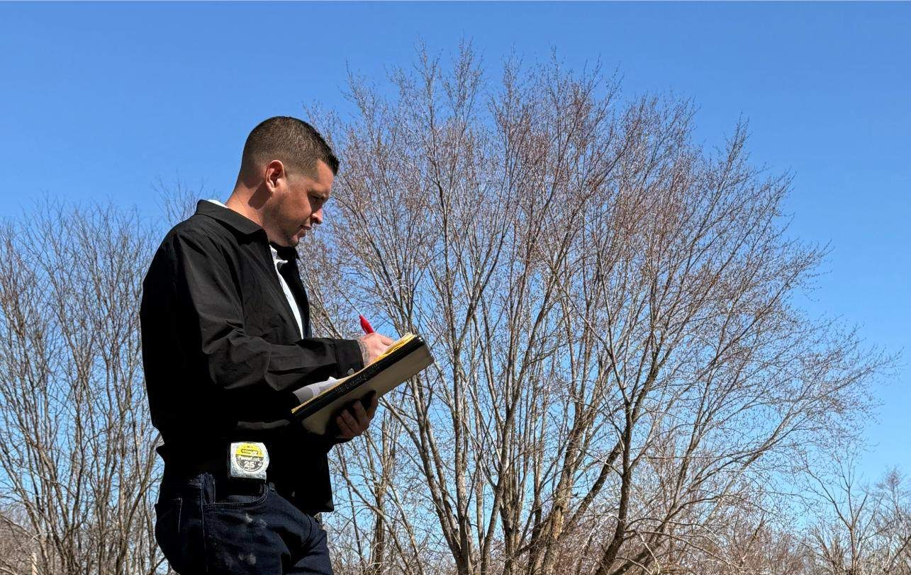 Person on a rooftop, taking notes. Sunny day. Trees in background, houses visible.