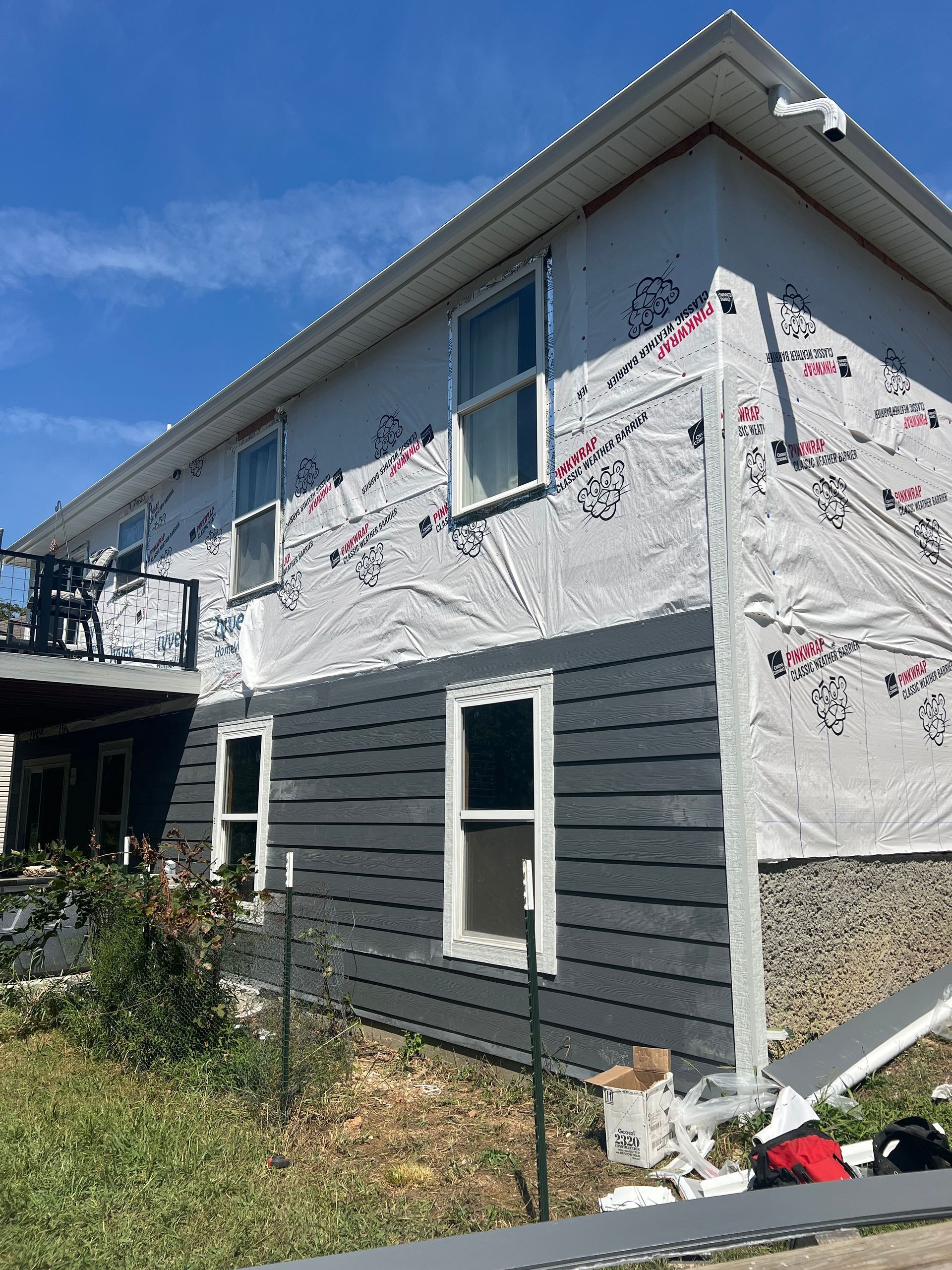 Two-story house with dark gray siding being installed, white wrap, blue sky.