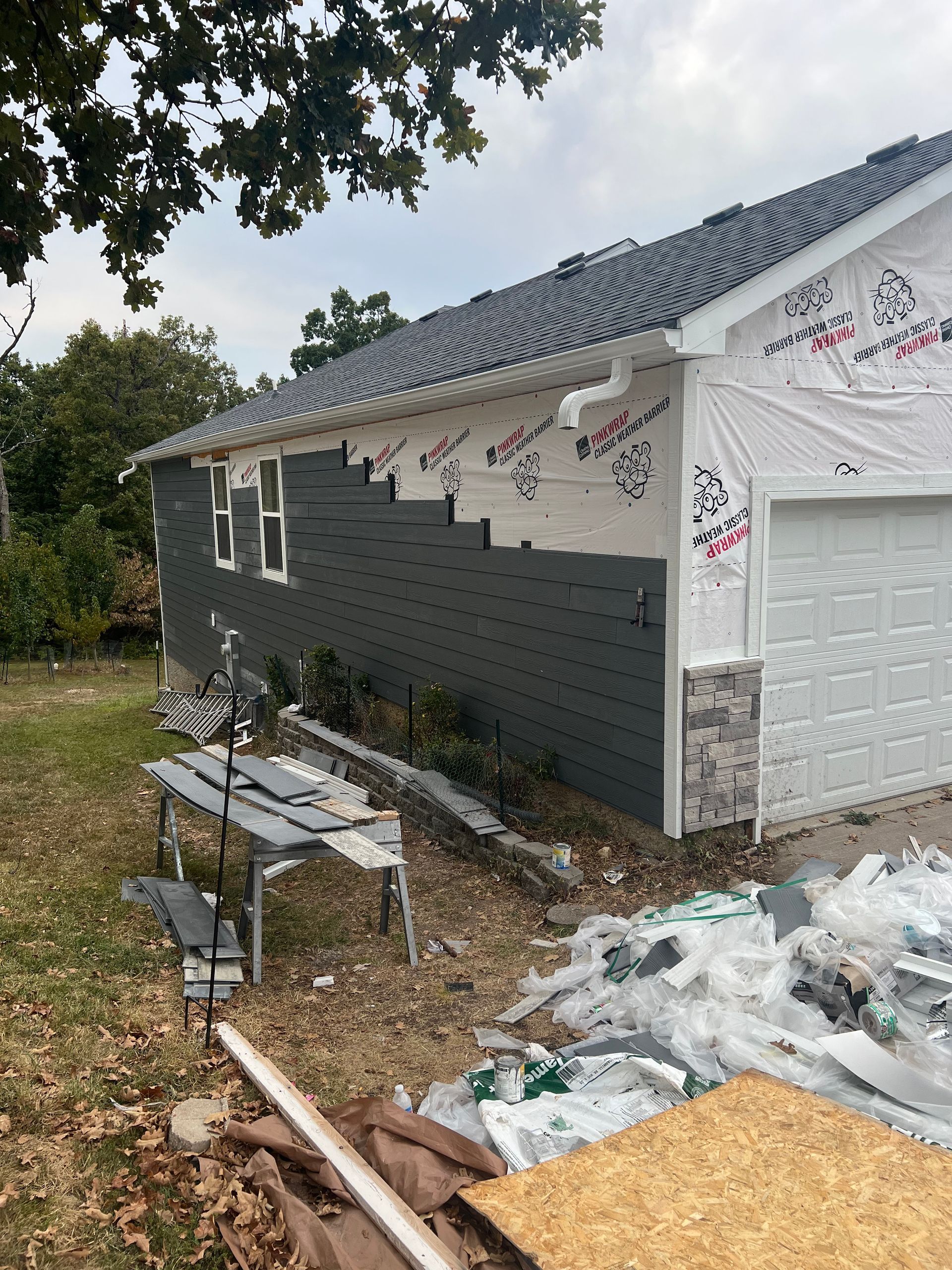 House exterior under construction, dark gray siding and stone facade, garage door, exposed wrap.