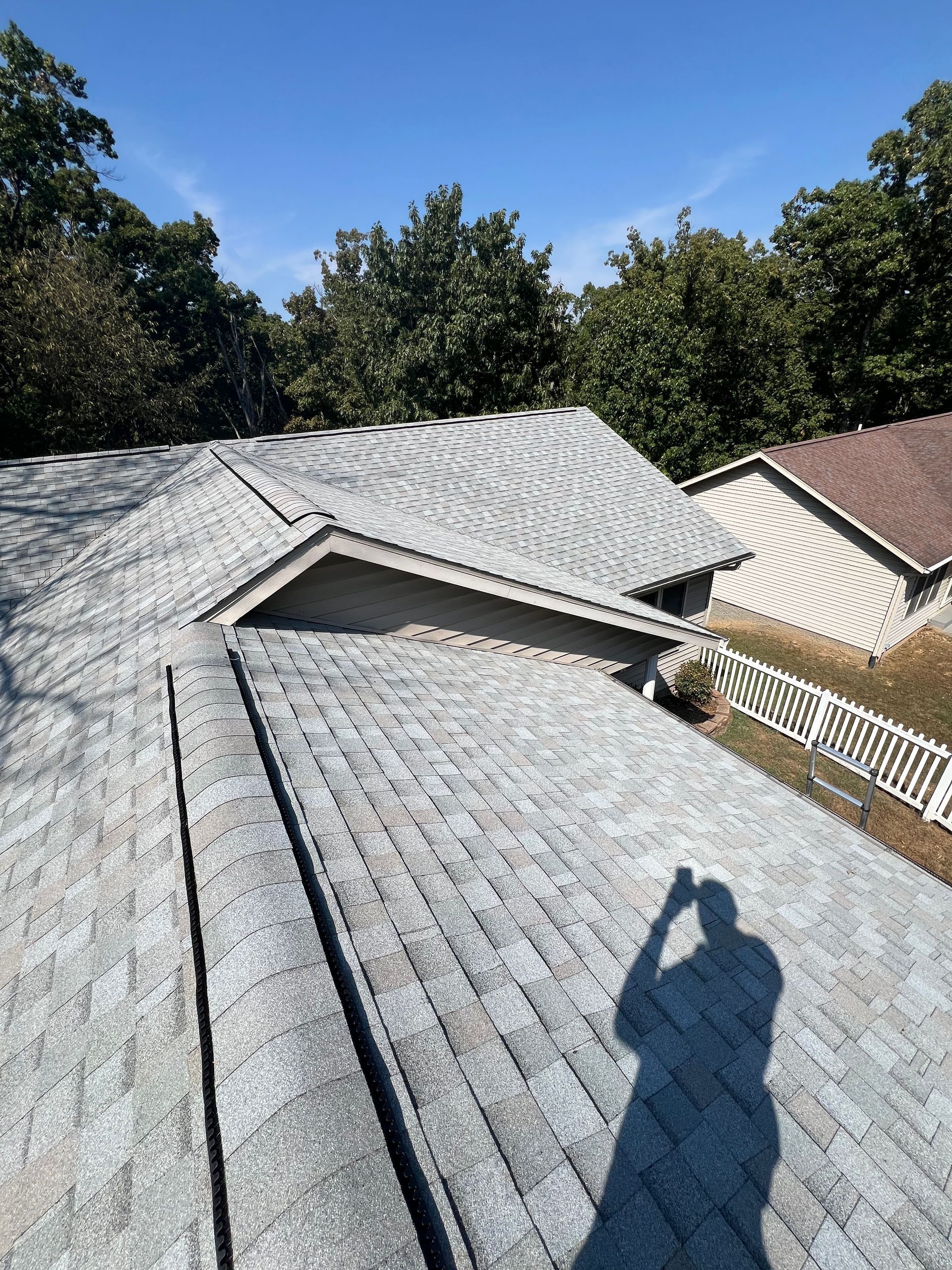 Gray shingled roof under a clear blue sky. Shadows of trees and a person taking a photo are visible.