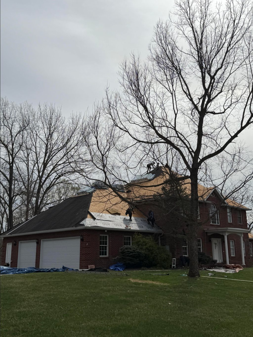 House with a partially torn-off roof under a cloudy sky, trees in the yard, and garage doors.