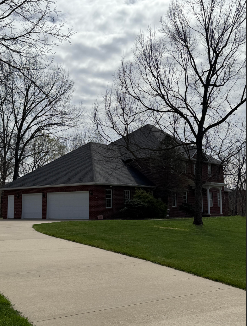 Red brick house with a three-car garage, surrounded by trees and a green lawn under a cloudy sky.