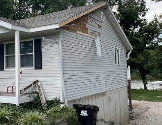 House with missing siding, revealing sheathing. Gray siding, white trim, with a black trash bin.
