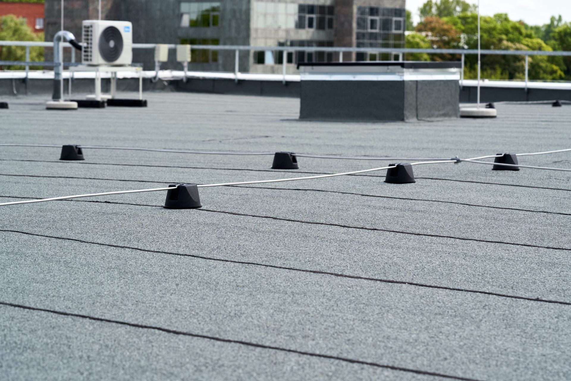 Flat, black, granular roof with black vents and an air conditioning unit; building background.
