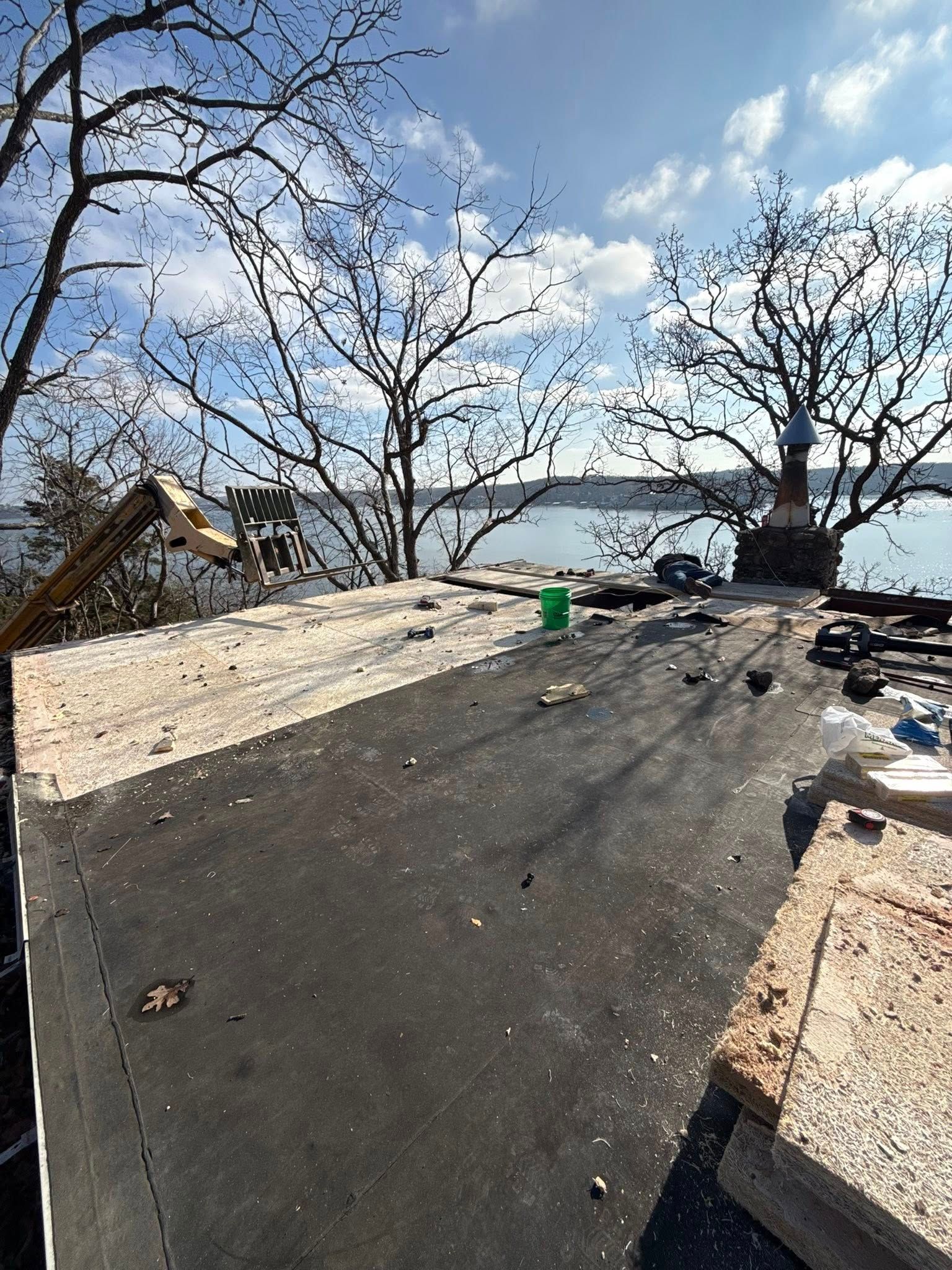 Rooftop view of a dark surface with construction debris; trees frame a water view under a cloudy sky.