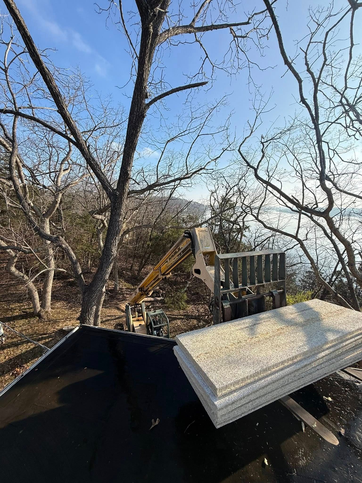 Construction site: A roof with insulation panels, a lift, and trees against a sunny sky and water.