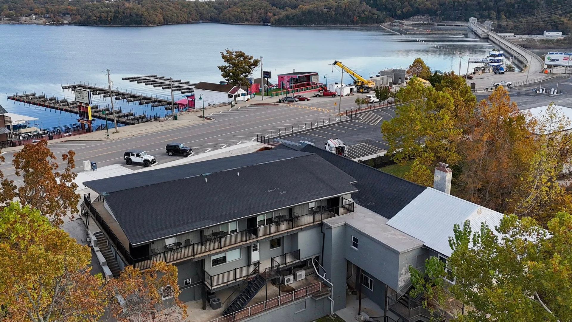 Overhead view of a waterfront building with a dark roof near docks and a lake; fall foliage present.