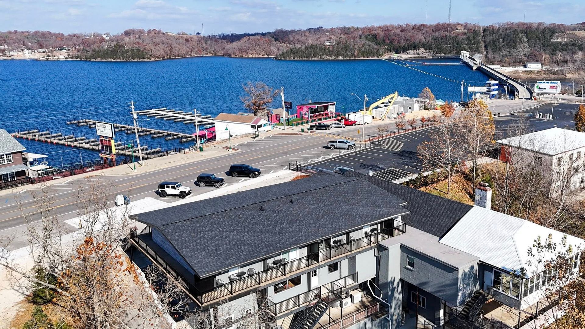 Aerial view of a lakefront area with buildings, road, marina, bridge, and clear blue water.