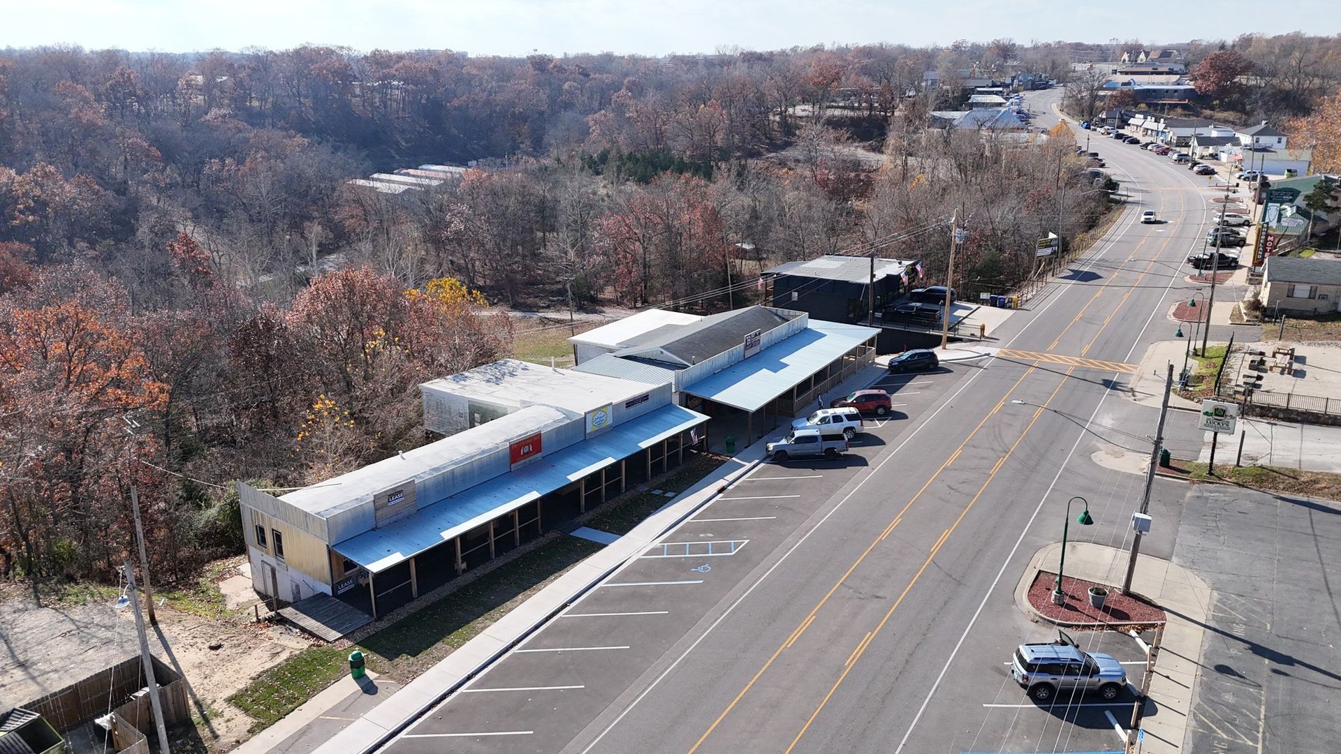 Aerial view of a long, light-colored building with a covered porch and parking lot, next to a street lined with other buildings and trees.