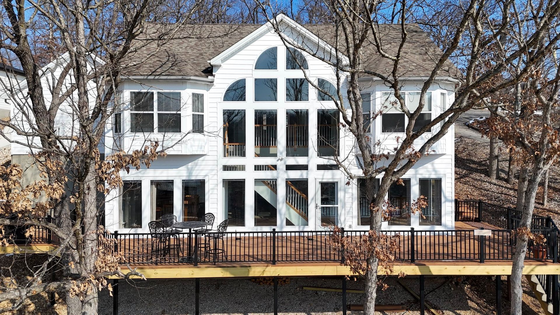 White house with large windows and a wooden deck, nestled among bare trees.
