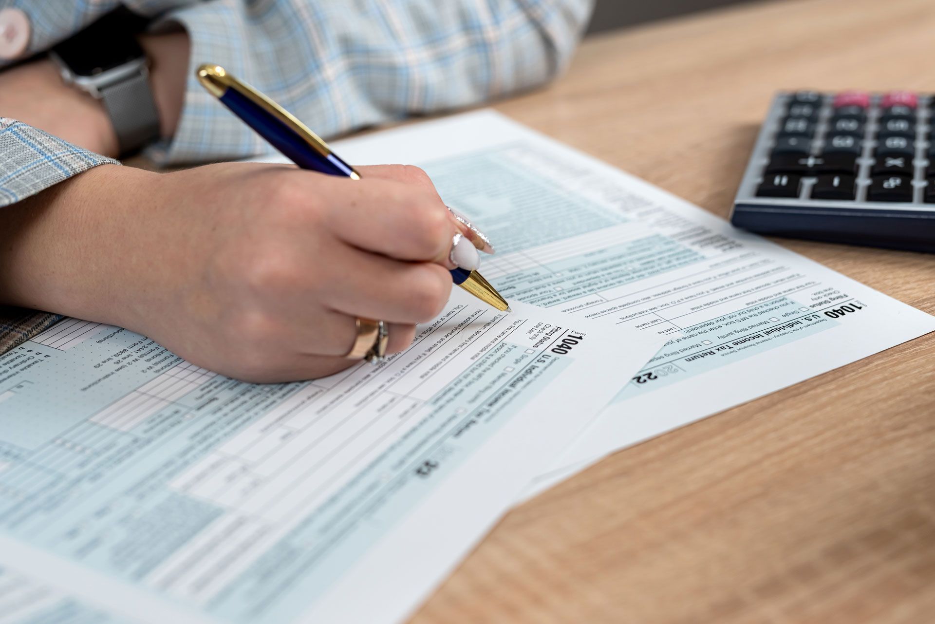 Person's hand writing on a tax form with a pen, next to a calculator on a wooden desk.