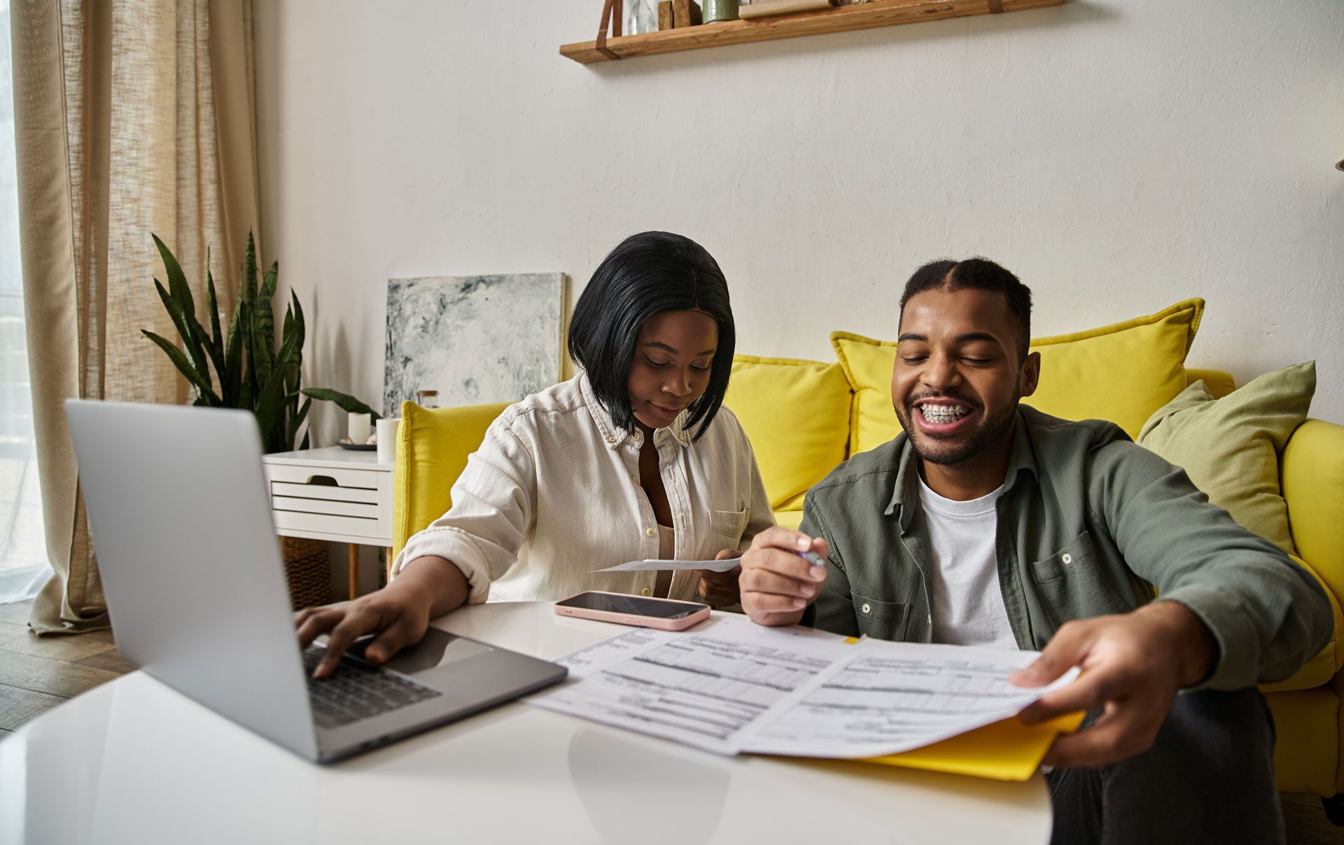Couple reviewing financial documents, using a laptop, smiling in a home setting.