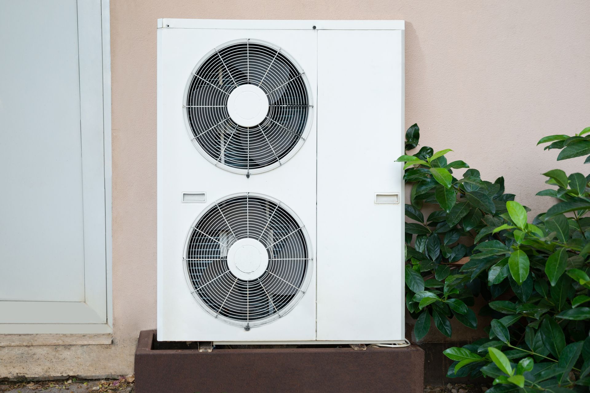 A white air conditioner is sitting outside of a building next to a bush.